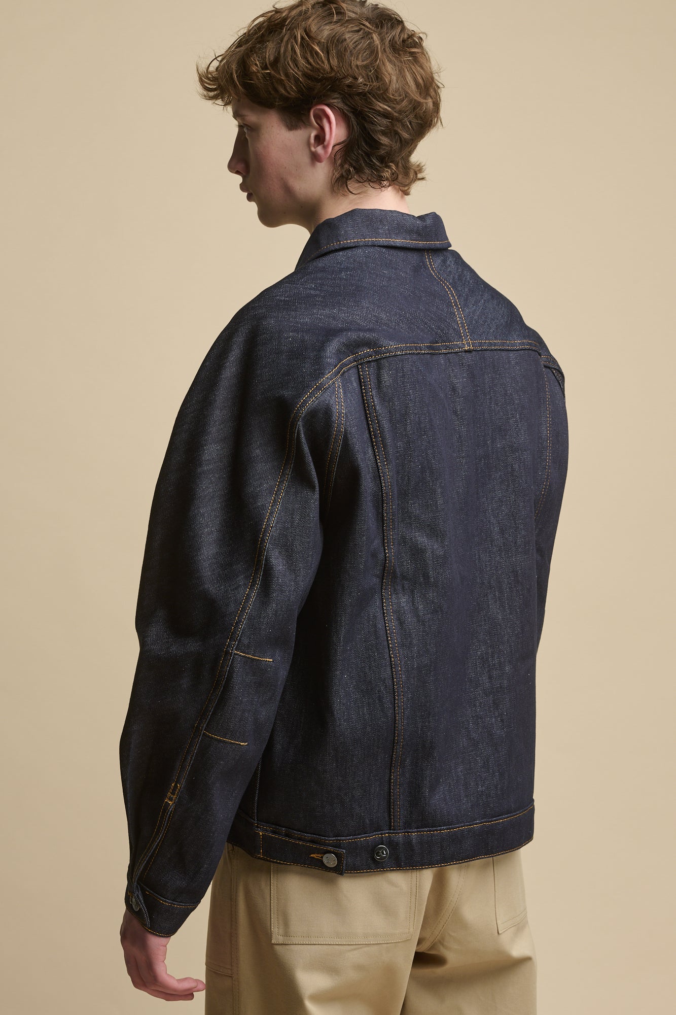 Thigh up image of male standing on an angle looking towards the camera wearing Joe Denim Jacket in indigo worn with raglan sweatshirt in cinnamon and cameraman pants in putty