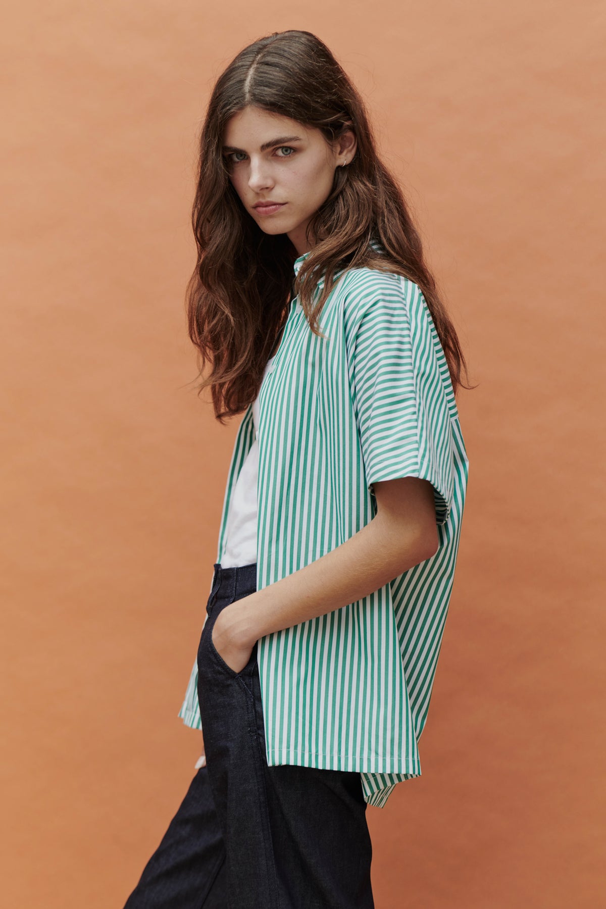 side angle shot of woman with long brown hair wearing a green and white short sleeved shirt with hands in her pockets