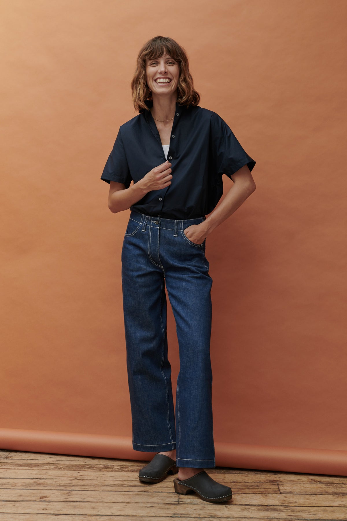 Front editorial short of woman in front oof Apricot background wearing the buttoned up Ava short sleeved shirt with clogs and wide fitting jeans