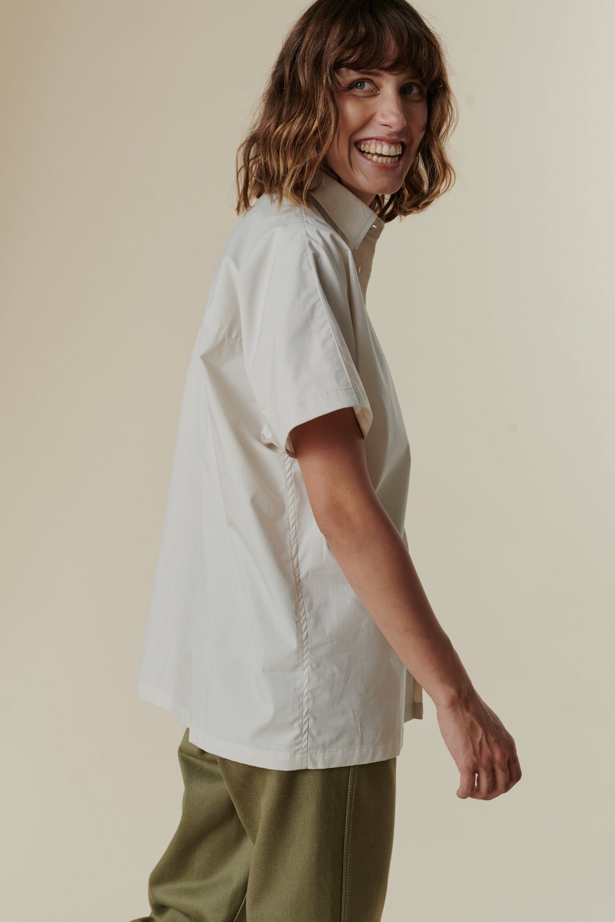Side angle of smiling woman with mid length brown hair wearing the short sleeve Ava shirt in white with olive green combat pants