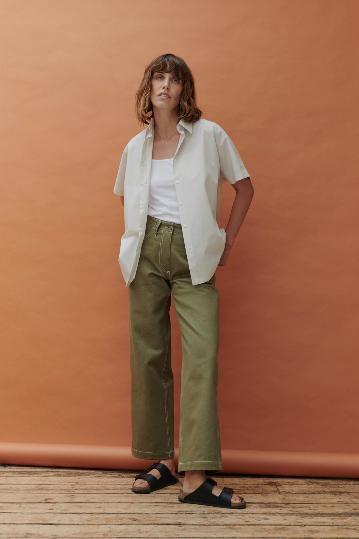 Woman with hands by her sides wearing the white Ava short sleeve shirt with a pale orange background