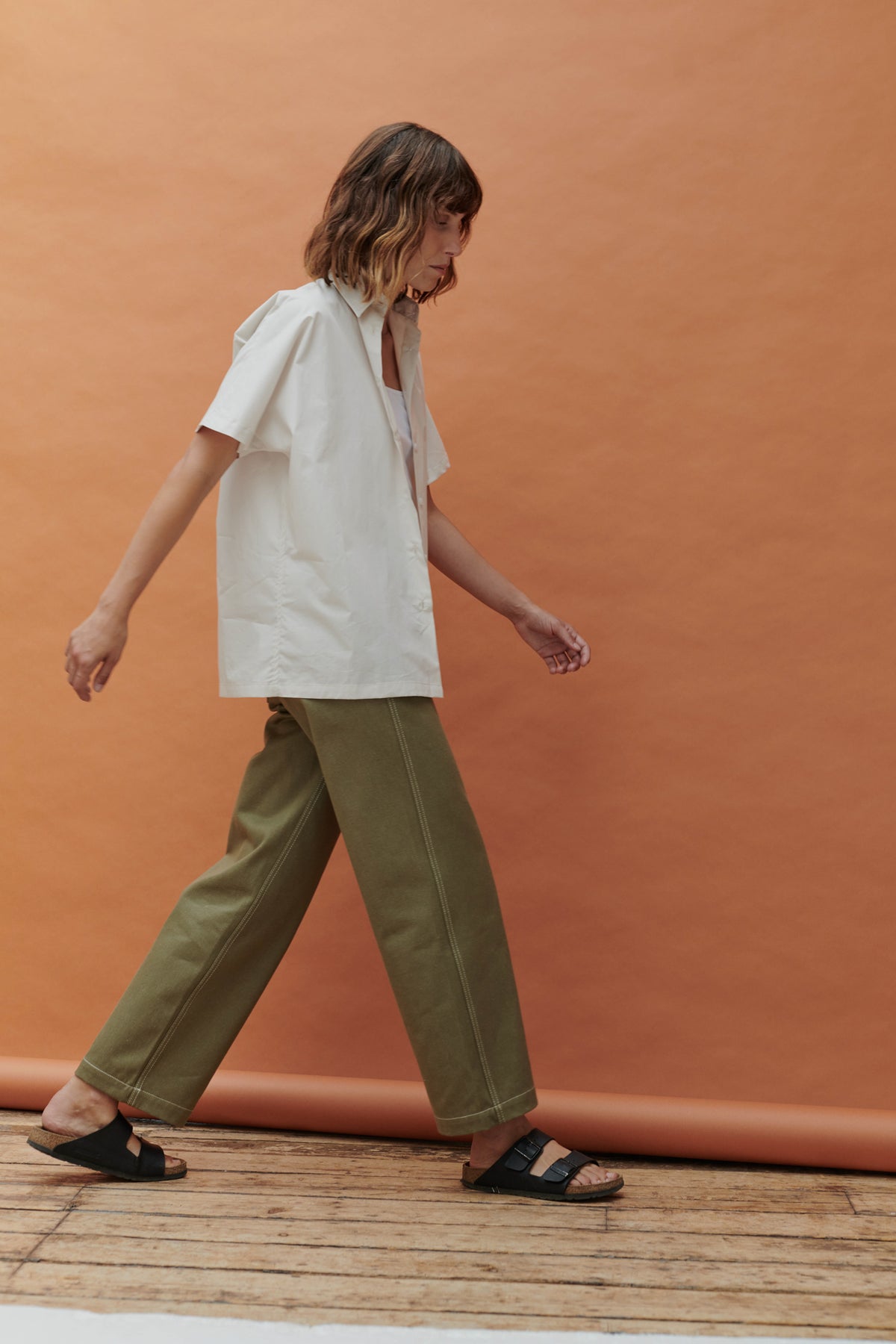Woman doing a walking pose infront of a pale orange background whilst wearing the short sleeve Ava shirt with the olive work jean