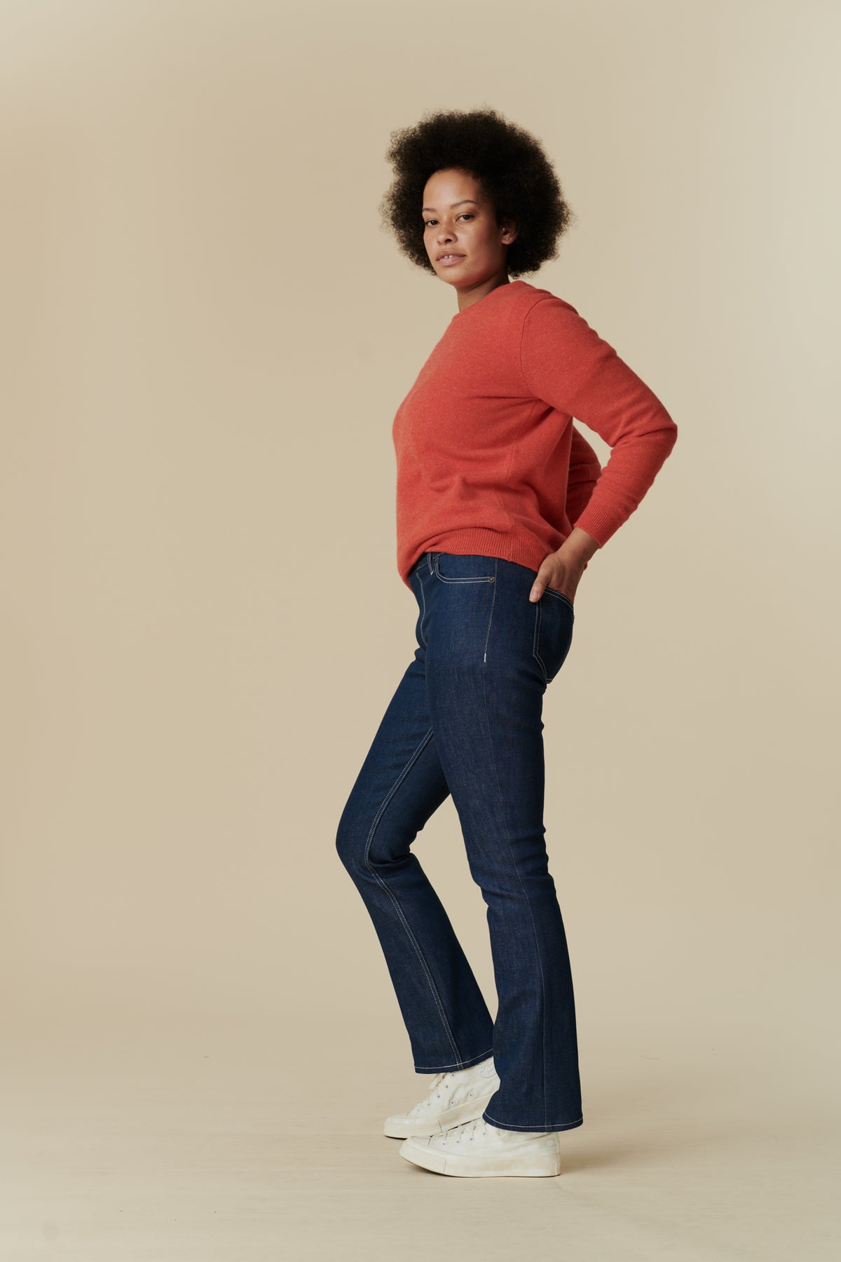 Full body side shot of black female model with afro stood with hands in back pockets of the women's bootcut mid rise jeans in blue paired with flame red, lambswool crew neck jumper