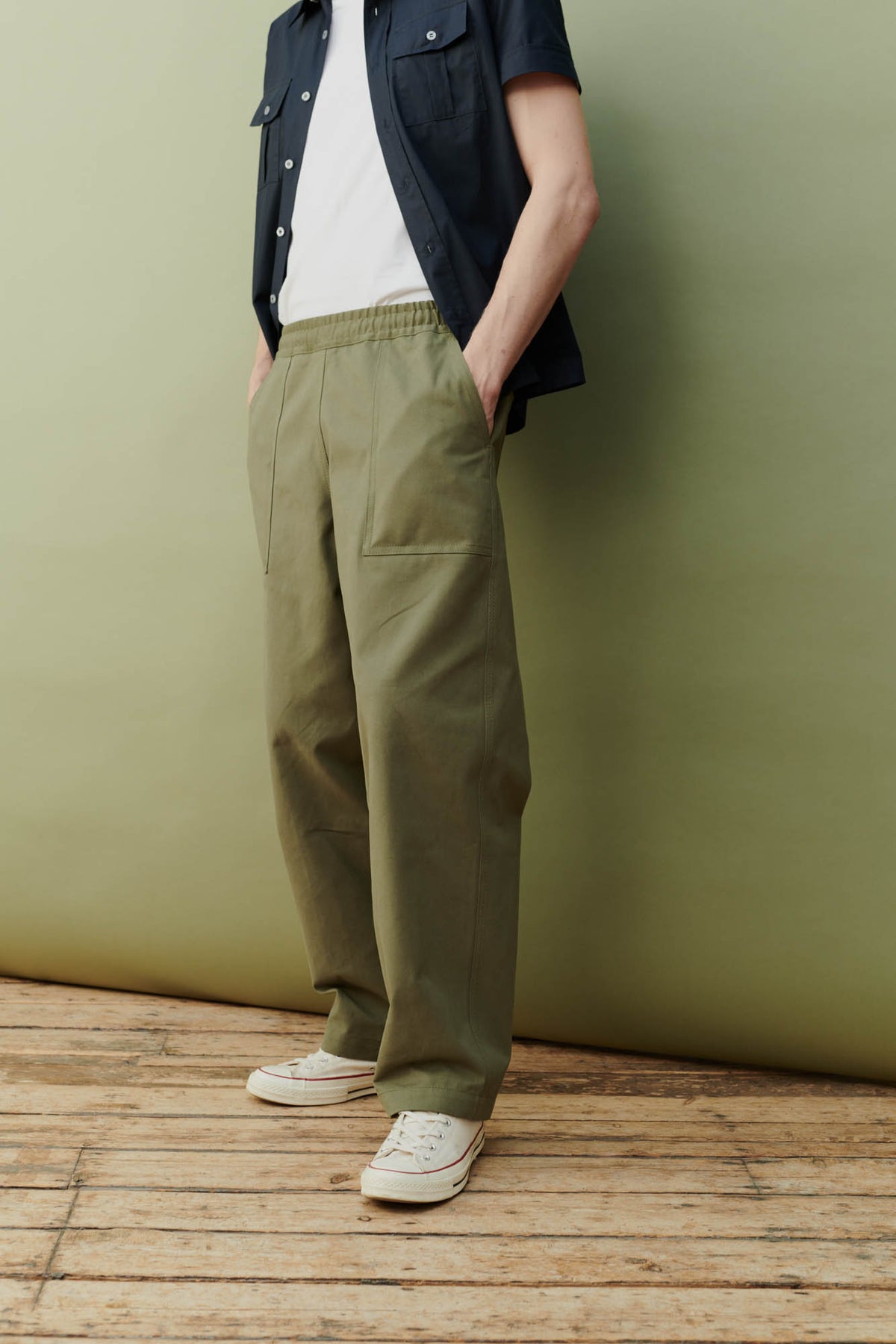 Side angle view of a man wearing olive green cameraman pant with a short sleeve navy shirt and a white t-shirt with white converse 70s trainers.