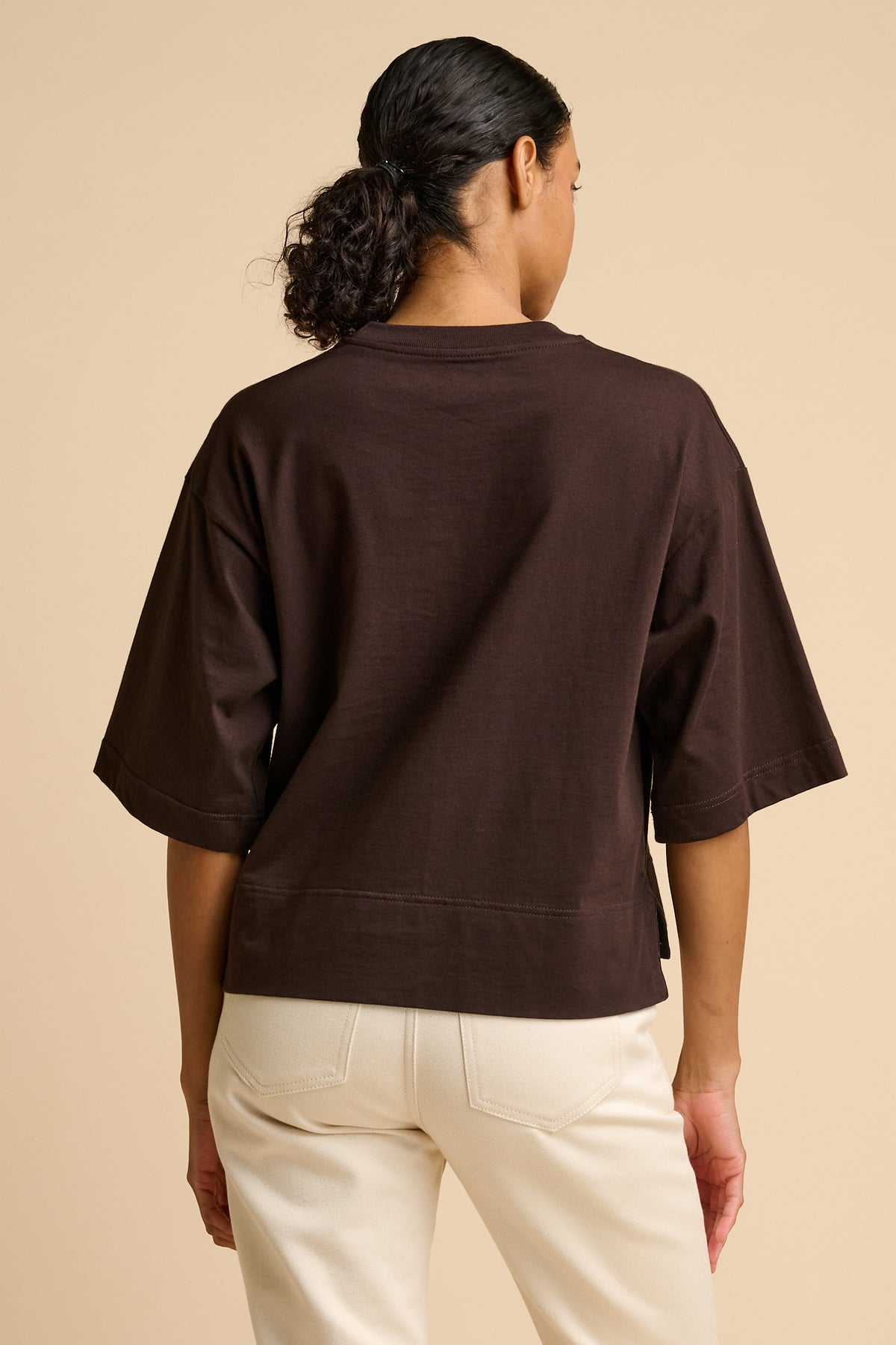 Woman wearing a brown t-shirt and white pants on a beige background.