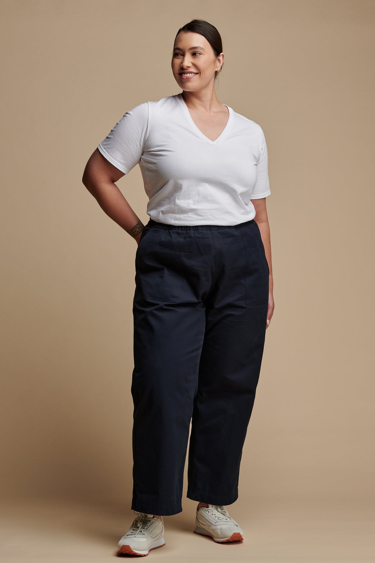 Full body of female wearing camerawoman drawstring tapered cotton canvas trousers in navy paired with v neck T shirt in white and Community Clothing X Walsh Parlick trainers.