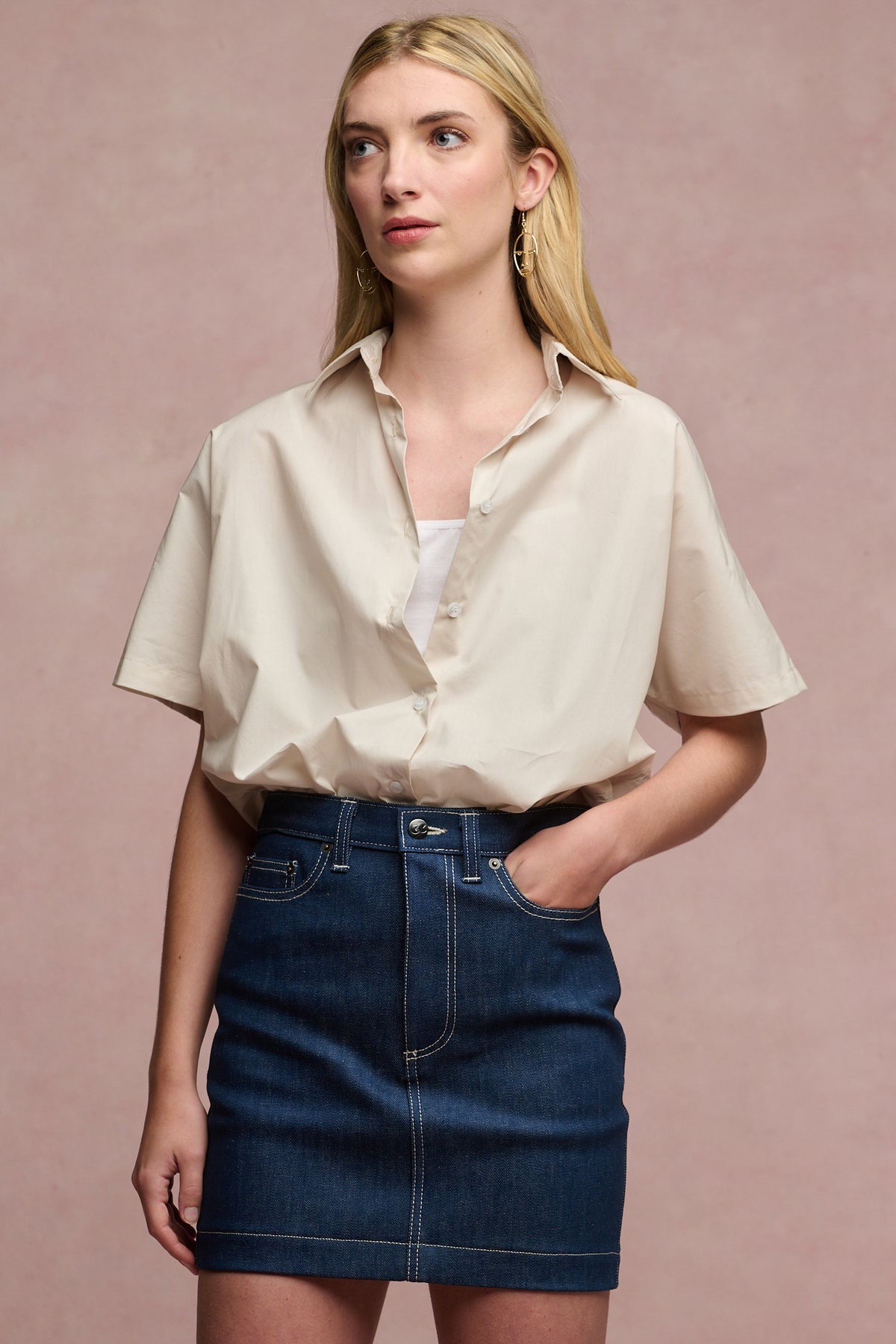 Thigh up image of blonde female wearing the Charlie denim skirt in blue, paired with camisole in white tucked in with Ava short sleeve shirt in stone