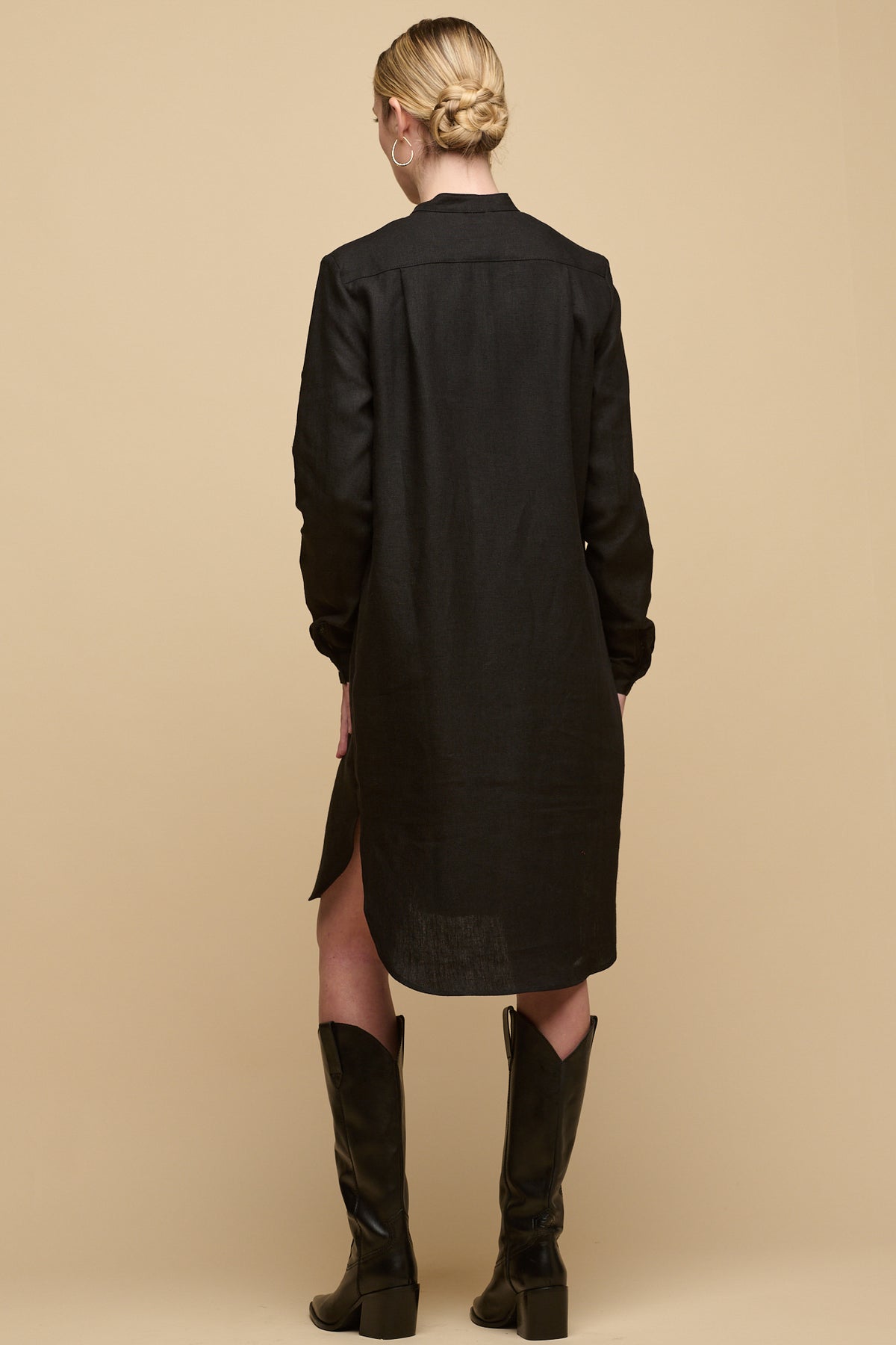 Full body image of the back of female wearing collarless shirt dress in black linen without belt for a looser fit paired with high boots