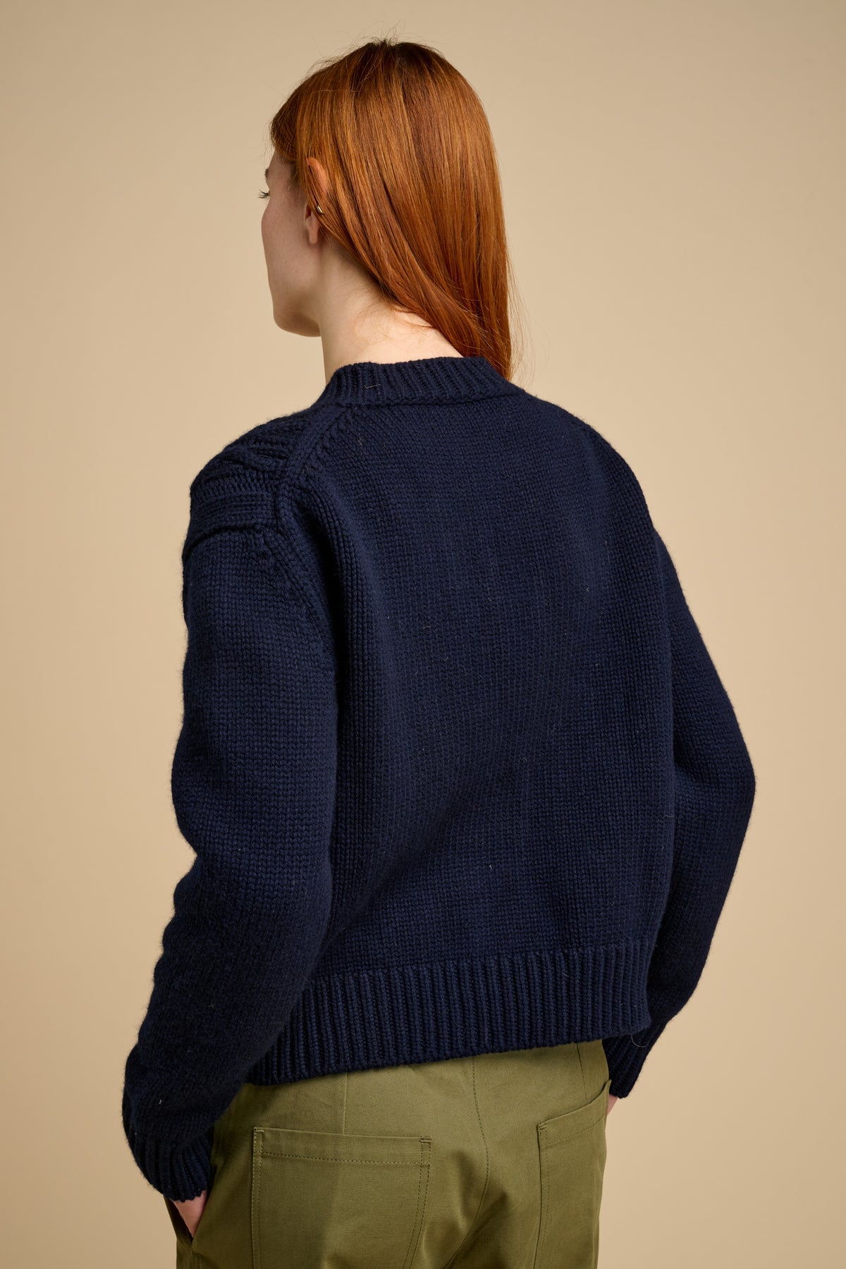 The back of female wearing a navy blue sweater and green pants on a beige background