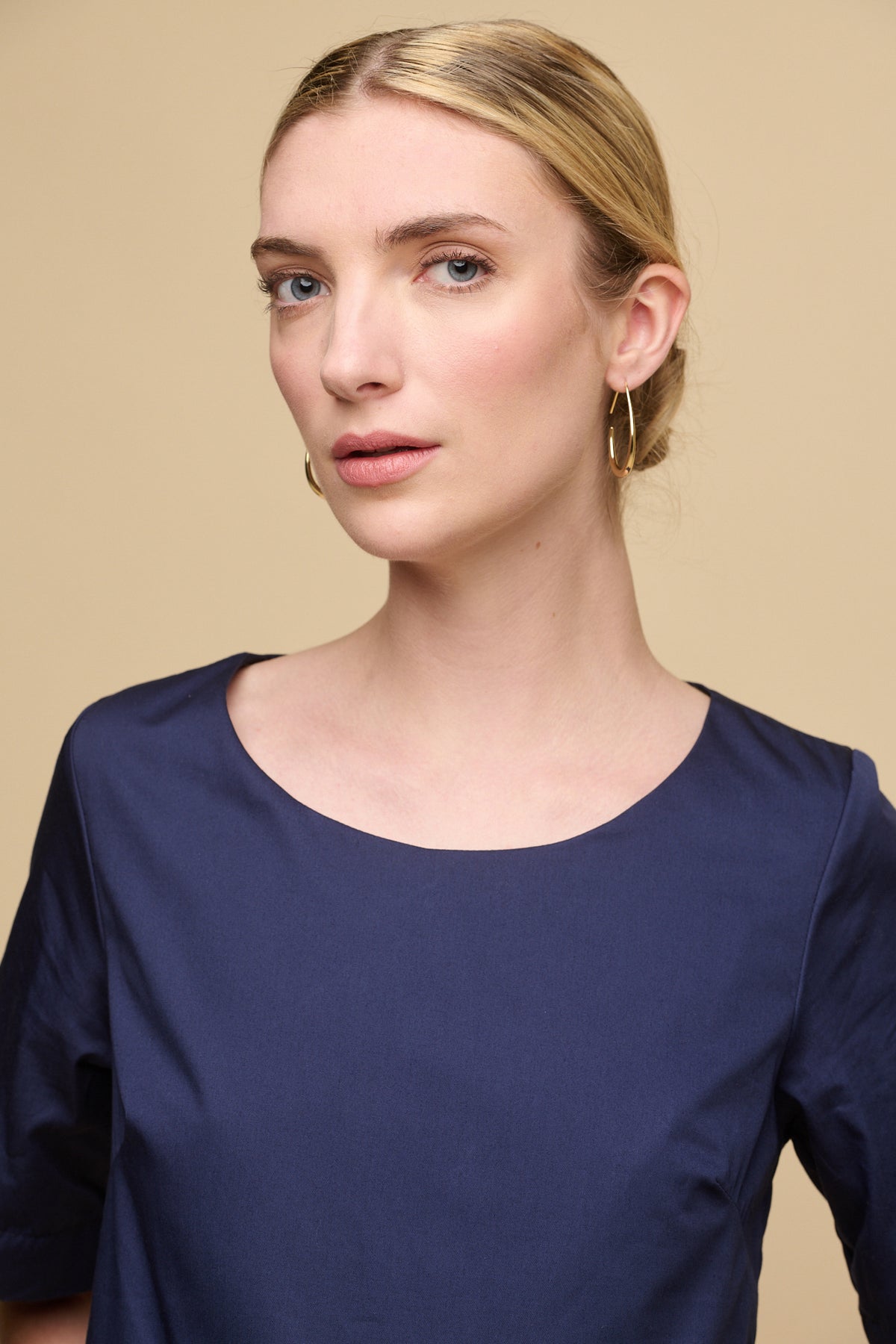 Portrait of female wearing crew neck gathered dress in navy with hair tied back in bun showing gold earrings