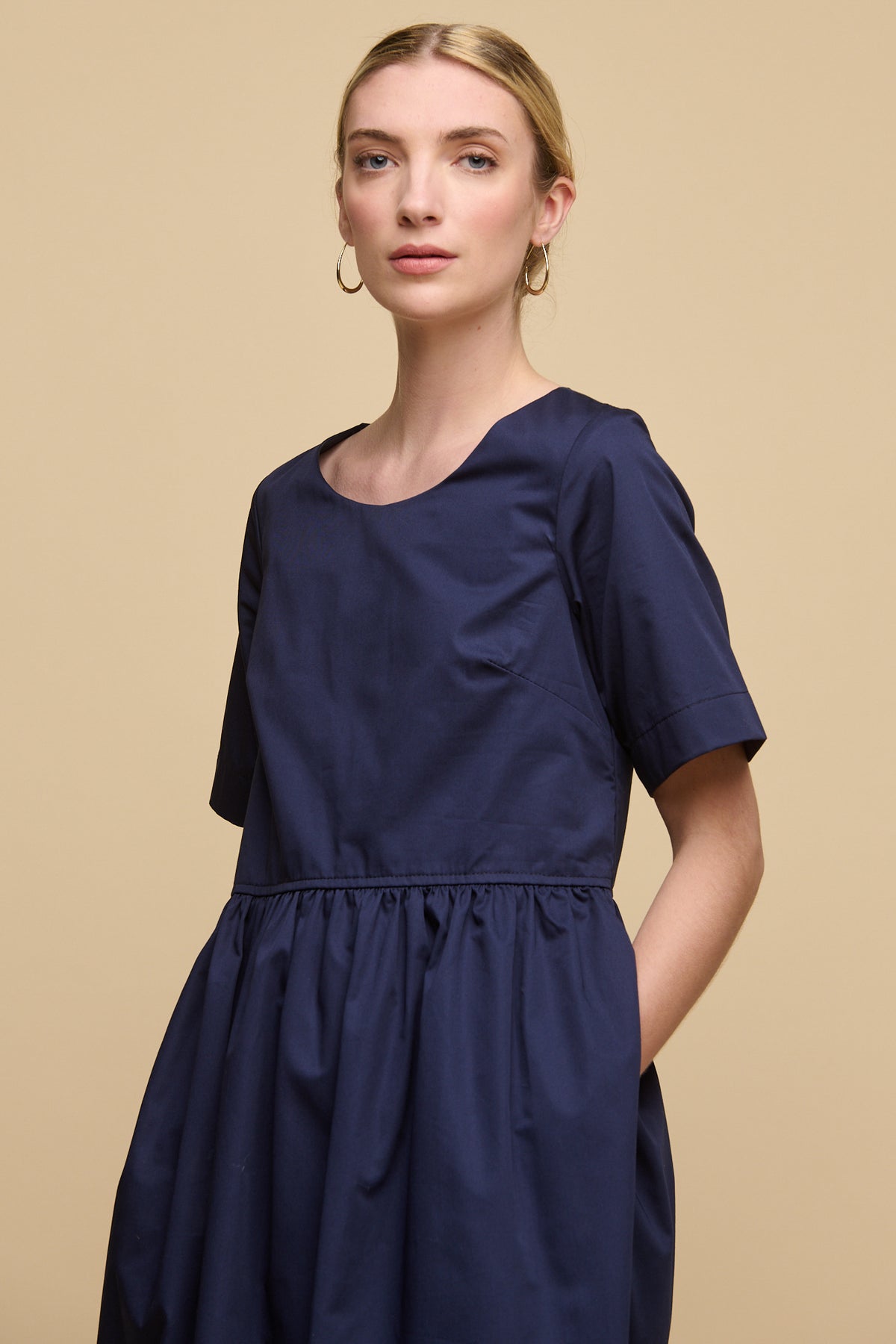 Thigh up image of blonde female with fair skin wearing crew neck gathered dress in navy with hands in pockets