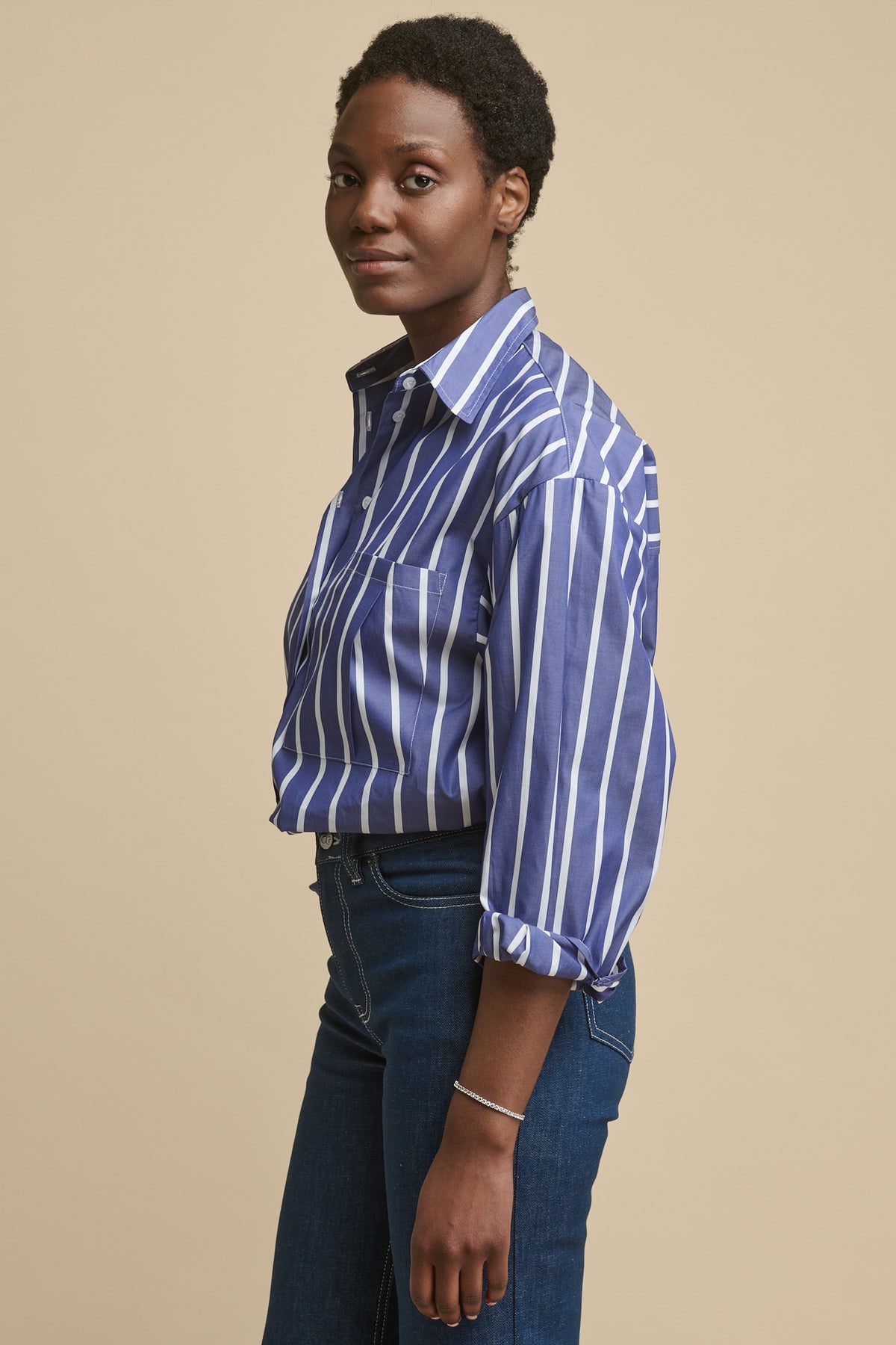 Thigh up image of the side of female wearing long sleeve Esme shirt in blue and white stripe tucked into blue jeans. Pleat detail patch pocket on chest and sleeves rolled up