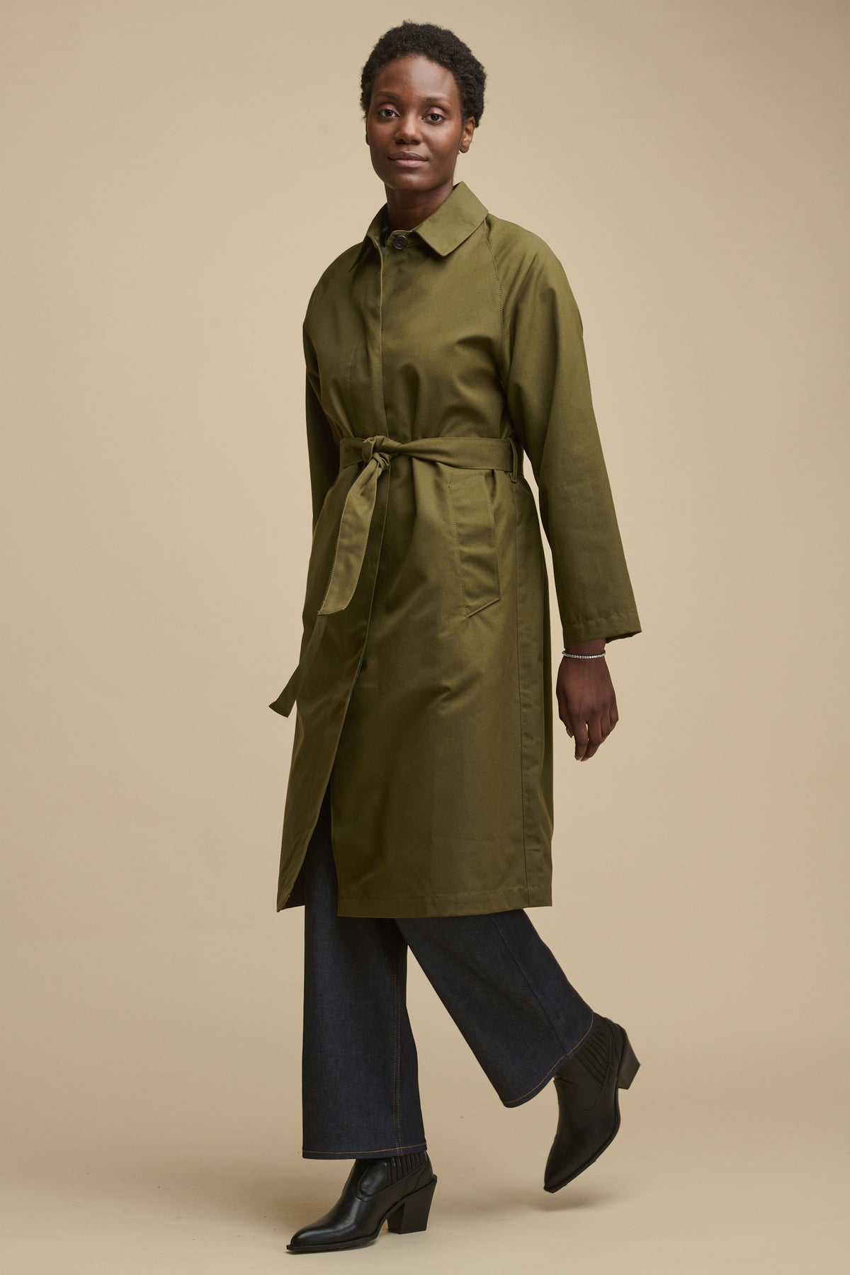 Full body image of black female facing the camera mid walk wearing Frances raglan belted raincoat with belt fastened over indigo jeans and black boots