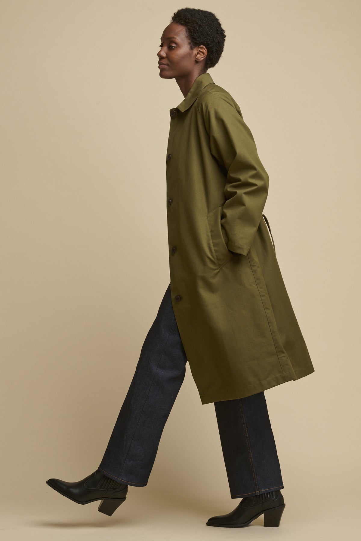Full body image of female from the side, mid walk wearing Frances Raglan Belted Raincoat in olive with indigo jeans and black boots