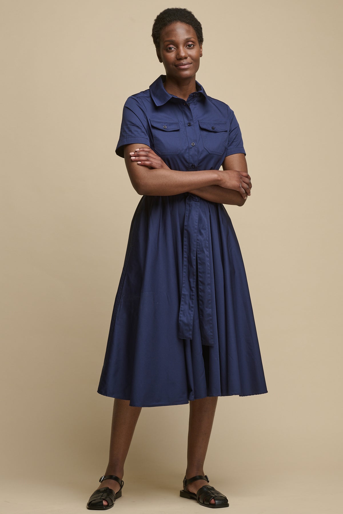 Full body image of the front of female wearing short sleeve shirt dress, with her arms folded over the waist belt tie, midi length dress