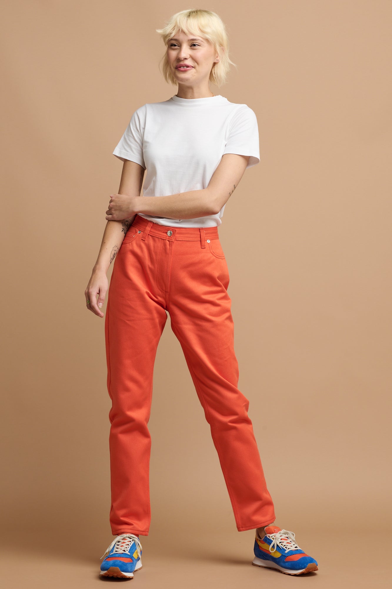 Female with short blonde hair wearing Community Clothing Gloria Tapered Jean in flame paired with white t shirt tucked in and Parlick trainers