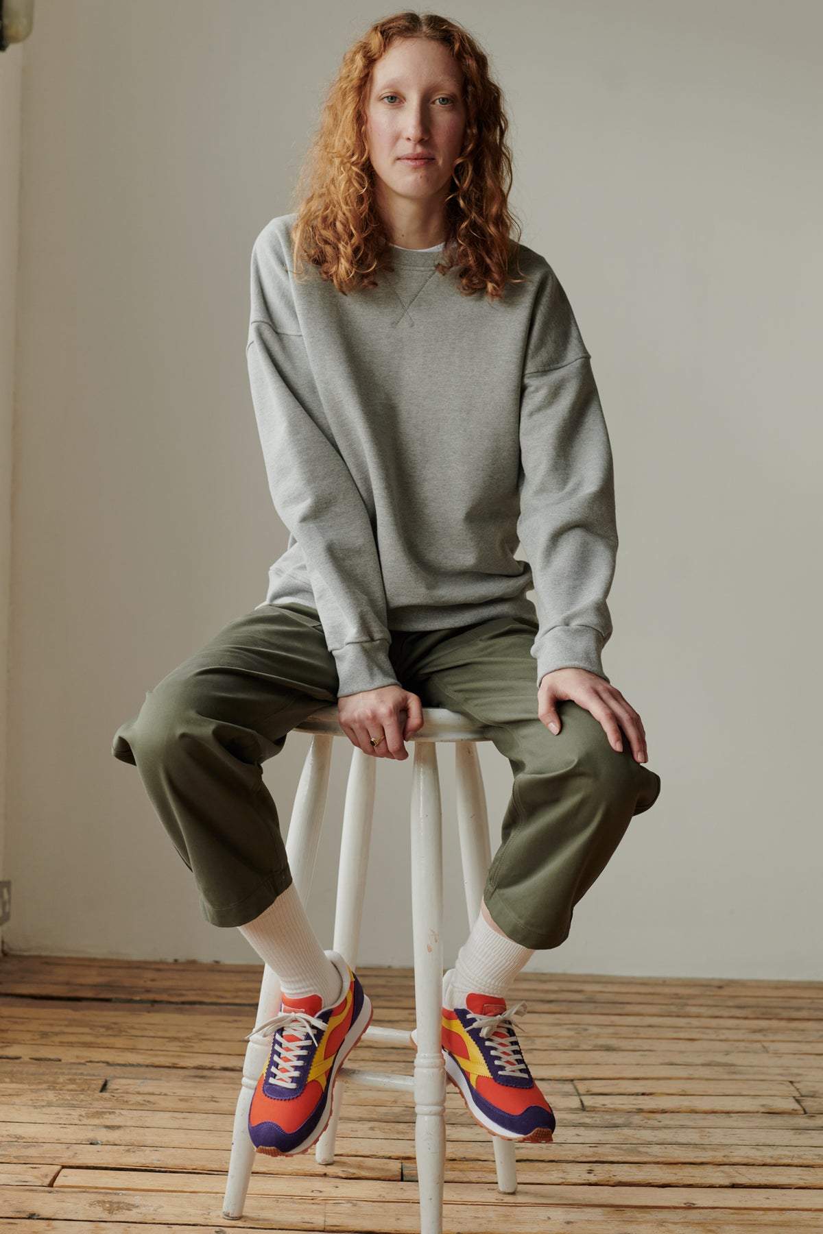 Curly haired ginger female wearing women's heritage sweatshirt in grey face on sat on tall stool 