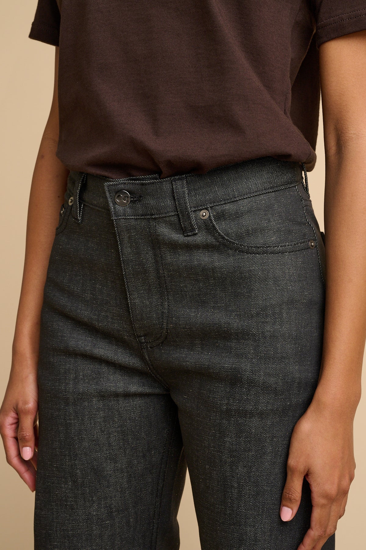 Woman wearing black jeans and a brown shirt against a beige background.