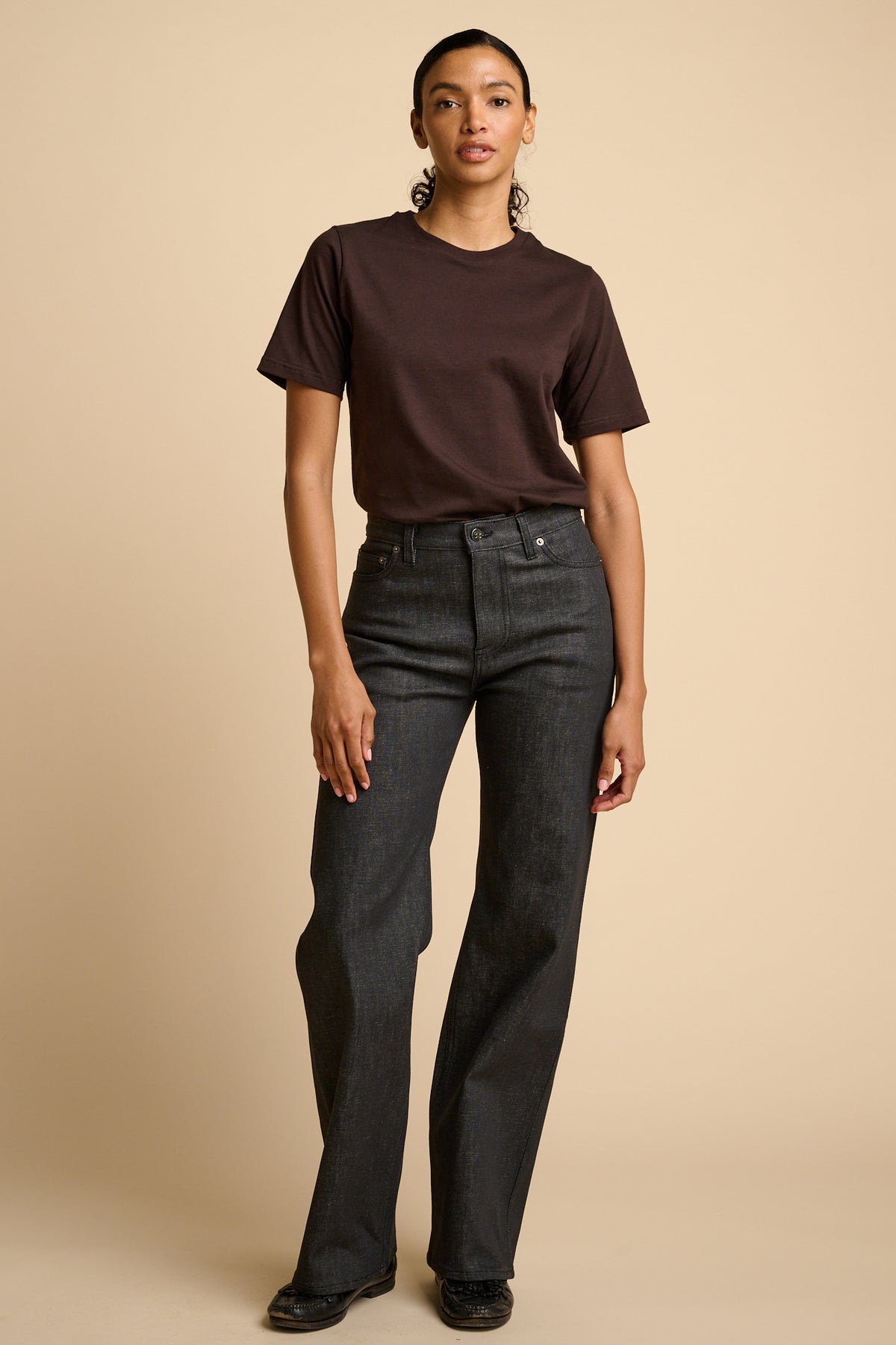 Woman wearing a brown t-shirt and black jeans on a beige background.