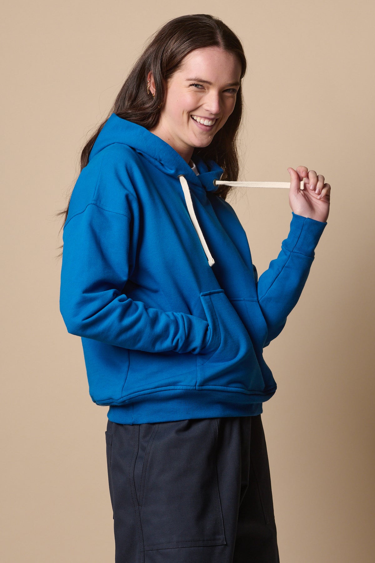 Smiley female wearing hooded sweatshirt in cobalt holding drawstring of the hood with one hand and one hand in the front pocket 