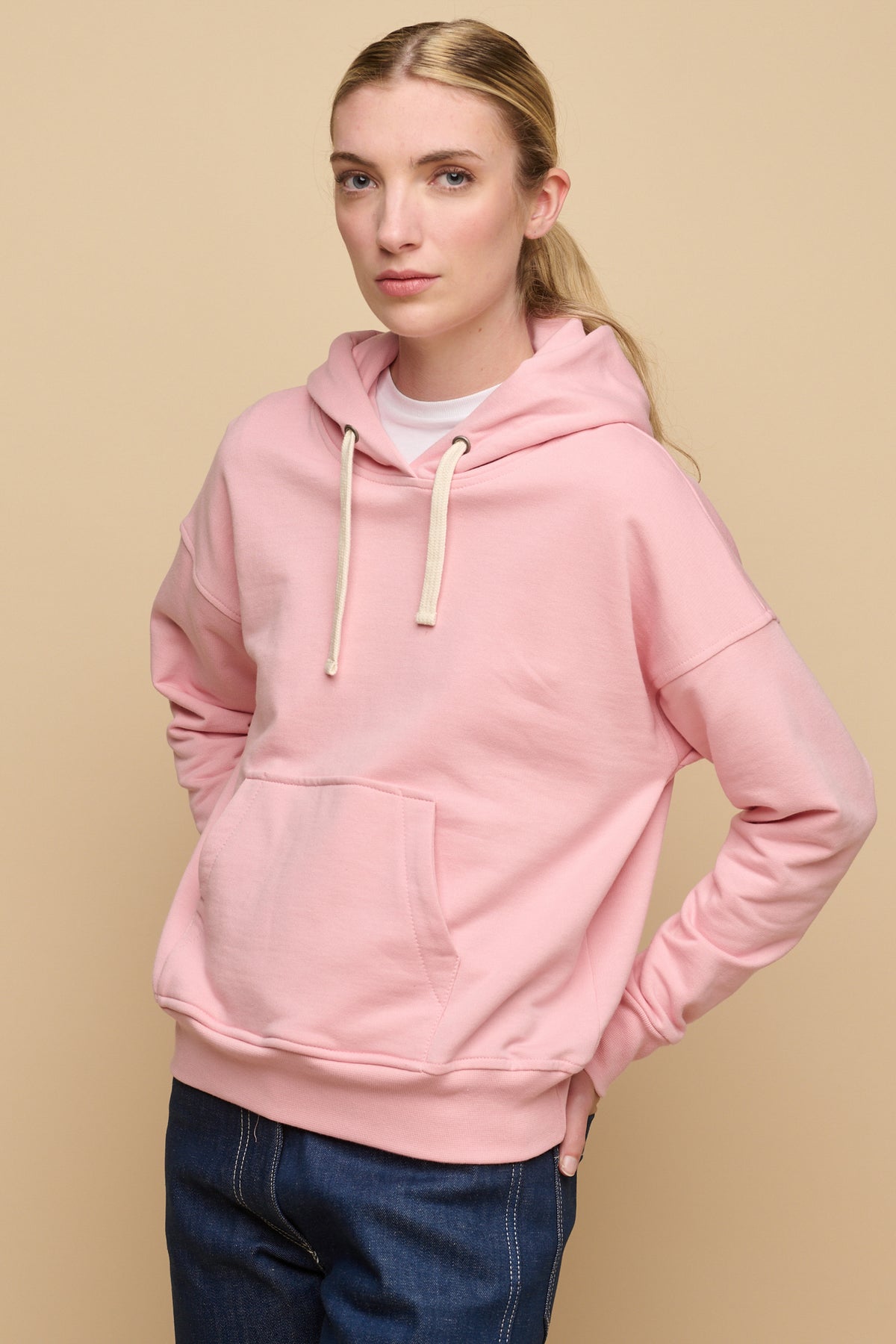 Thigh up image of the  front of female wearing pale pink hooded sweatshirt paired with work jeans in blue