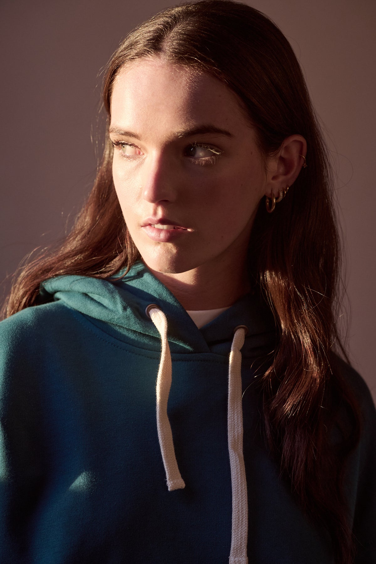 Portrait of female with long brown hair and sun shining on her face creating a shadow on the other side, wearing hooded sweatshirt in teal. 