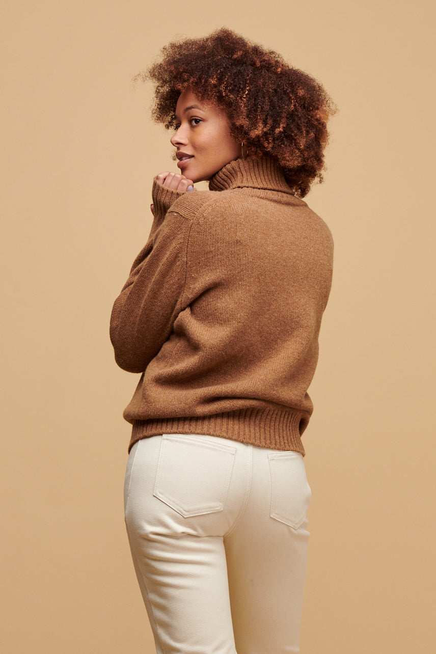 Thigh up of smiley black female with an afro wearing lambswool roll neck in dark camel paired with Community Clothing Gloria jeans in ecru