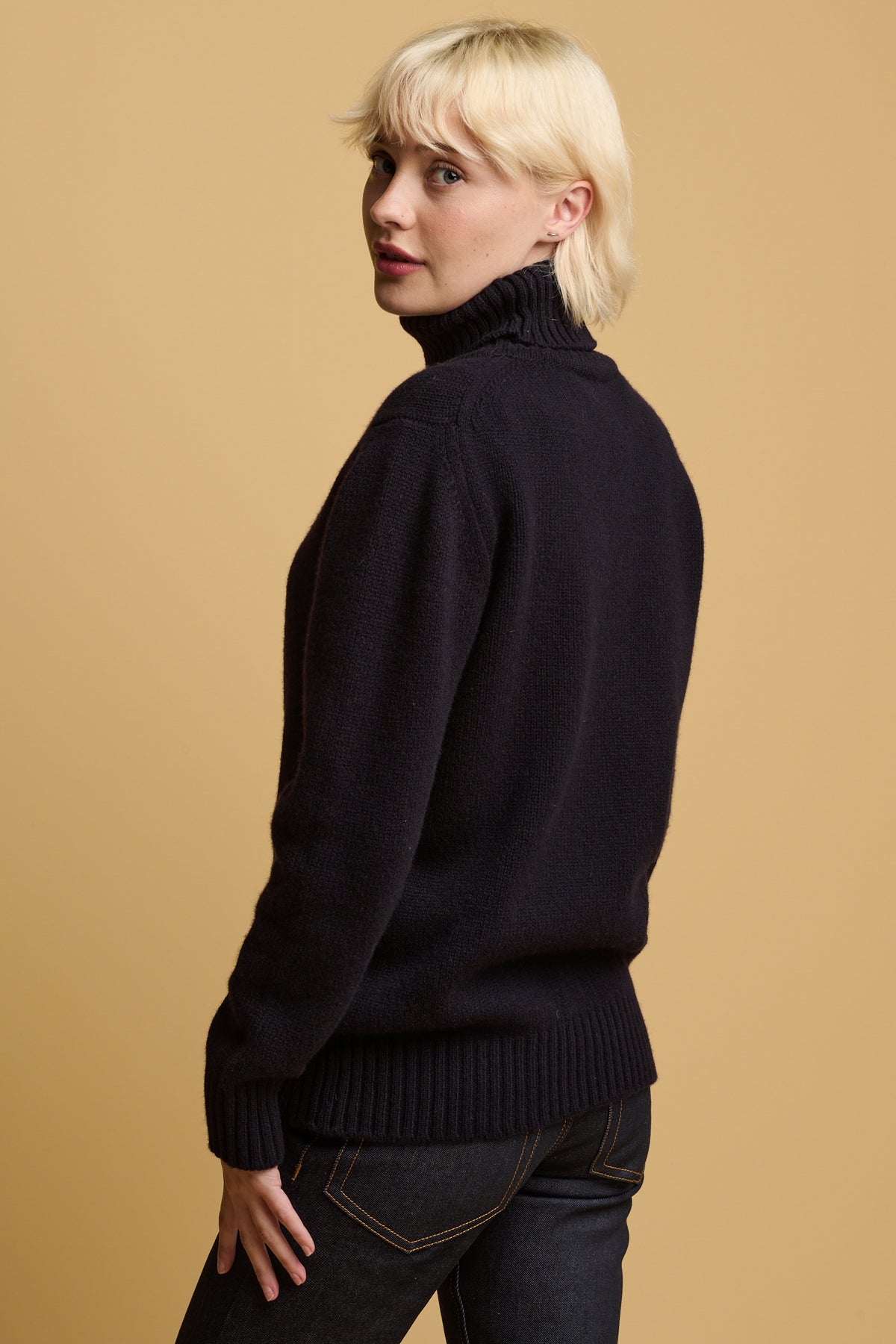 Thigh up image of the back of female looking over her shoulder back to the camera wearing lambswool roll neck jumper in navy