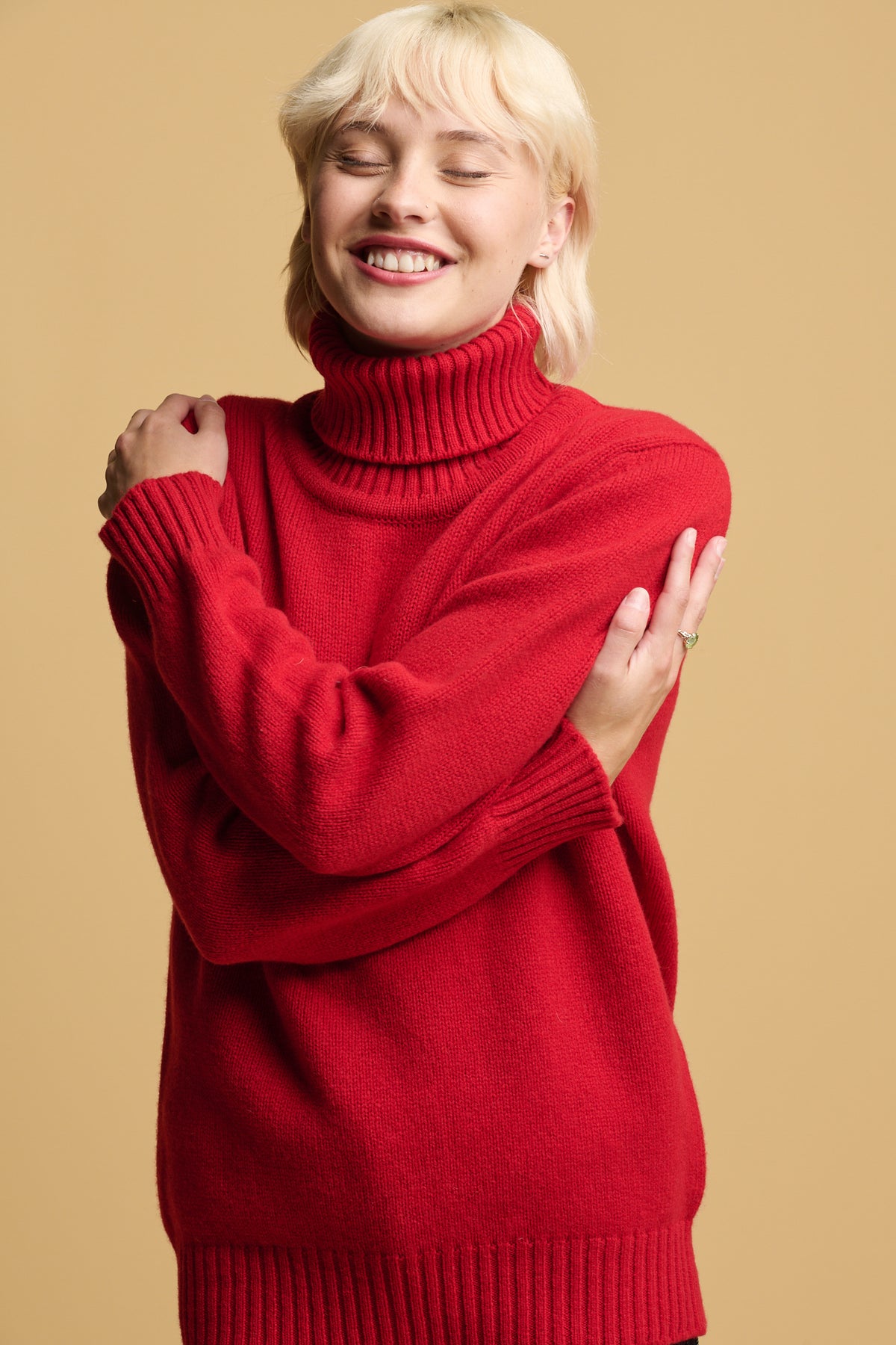 Smiley female with blond hair hugging herself in her soft cosy community clothing lambswool knit roll neck jumper in red