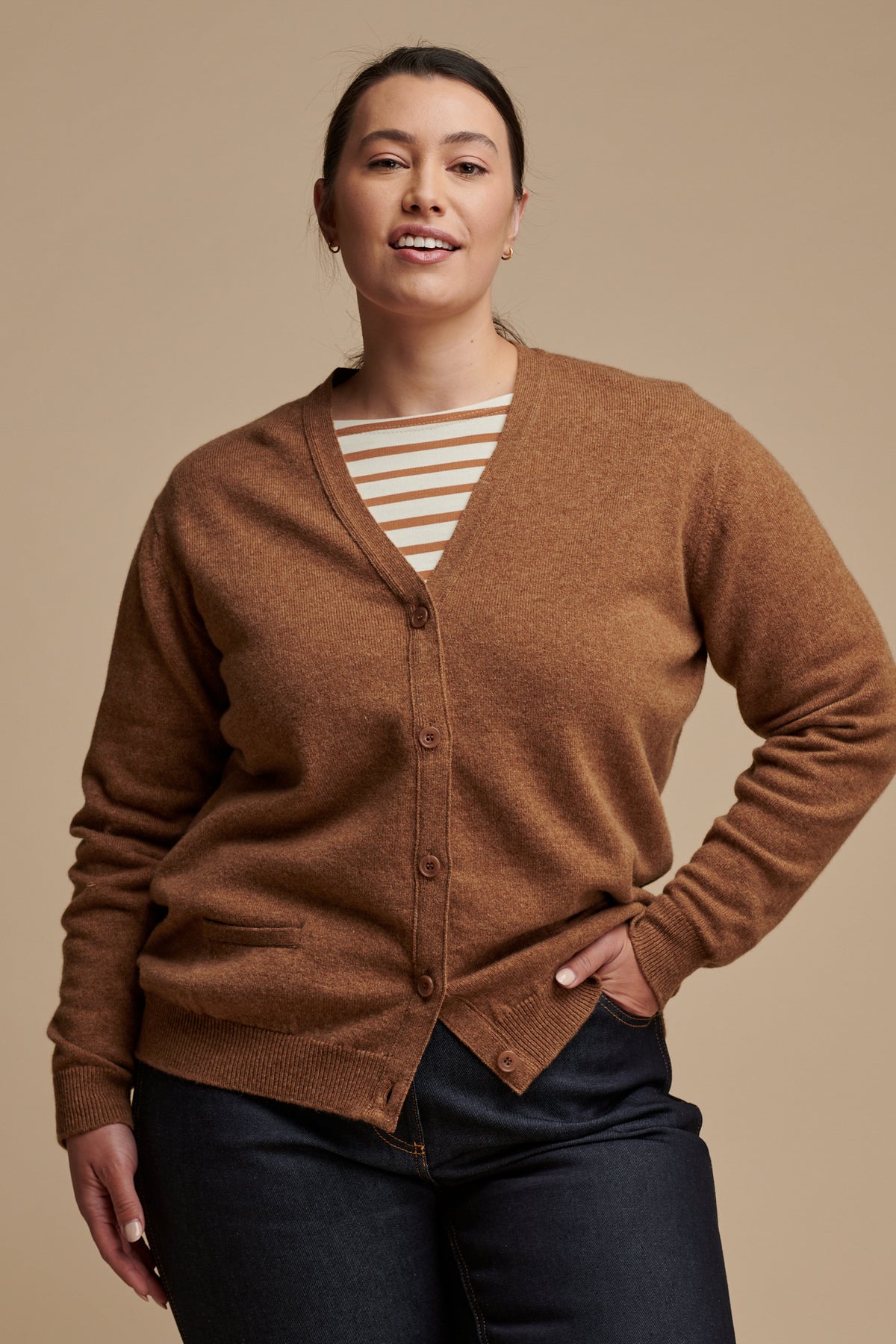 Thigh up image of female with her hand in the front jeans pocket wearing lambswool v neck cardigan in dark camel buttoned up over Breton top in ecru-cinnamon, paired with indigo jeans.