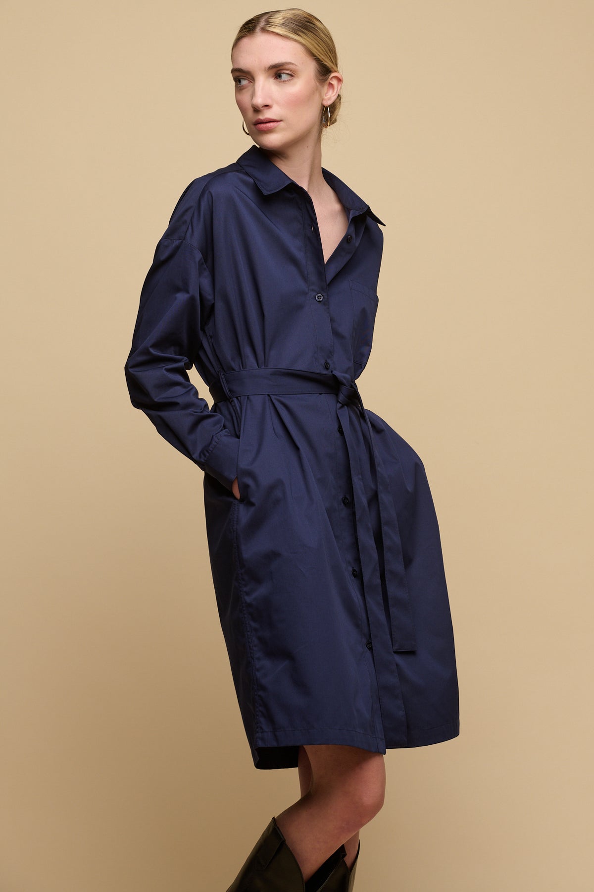 Blonde model with hair tied up wearing lorelle belted midi cotton shirt dress in navy with hands in pockets.