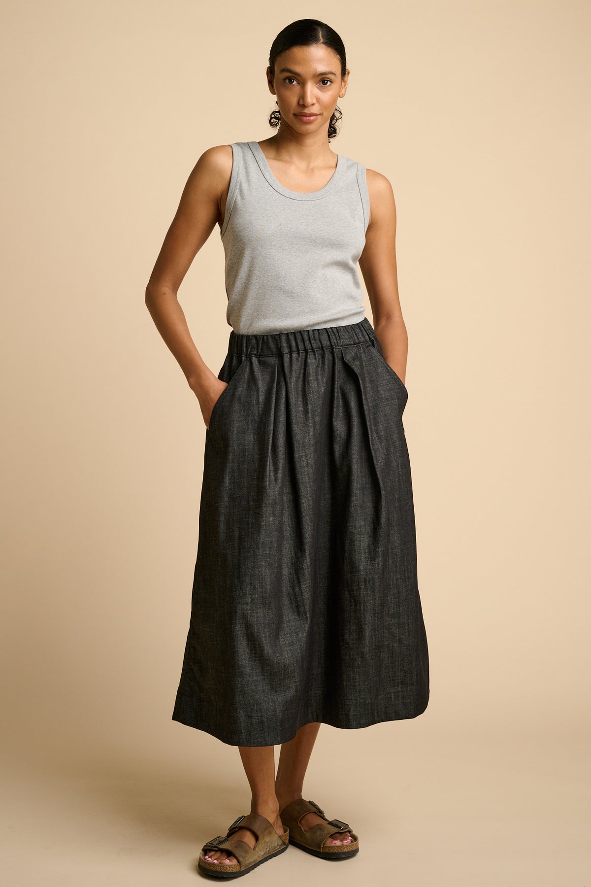 Woman wearing a grey tank top and black skirt on a beige background.