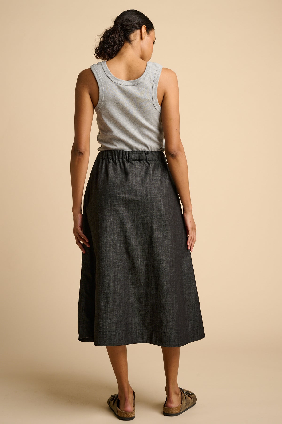 Woman wearing a grey tank top and black skirt on a beige background.