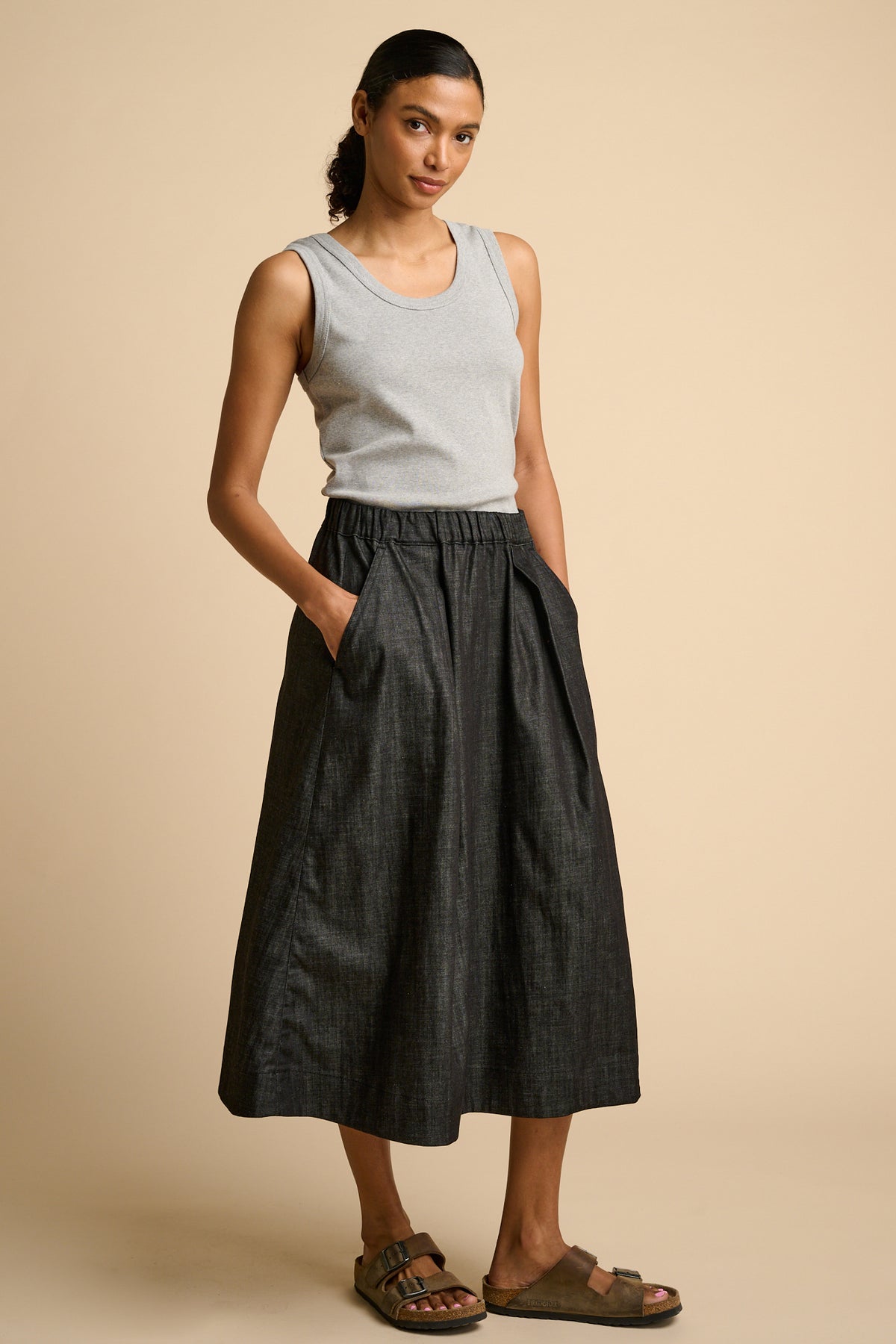 Woman wearing a grey tank top and black skirt on a beige background.