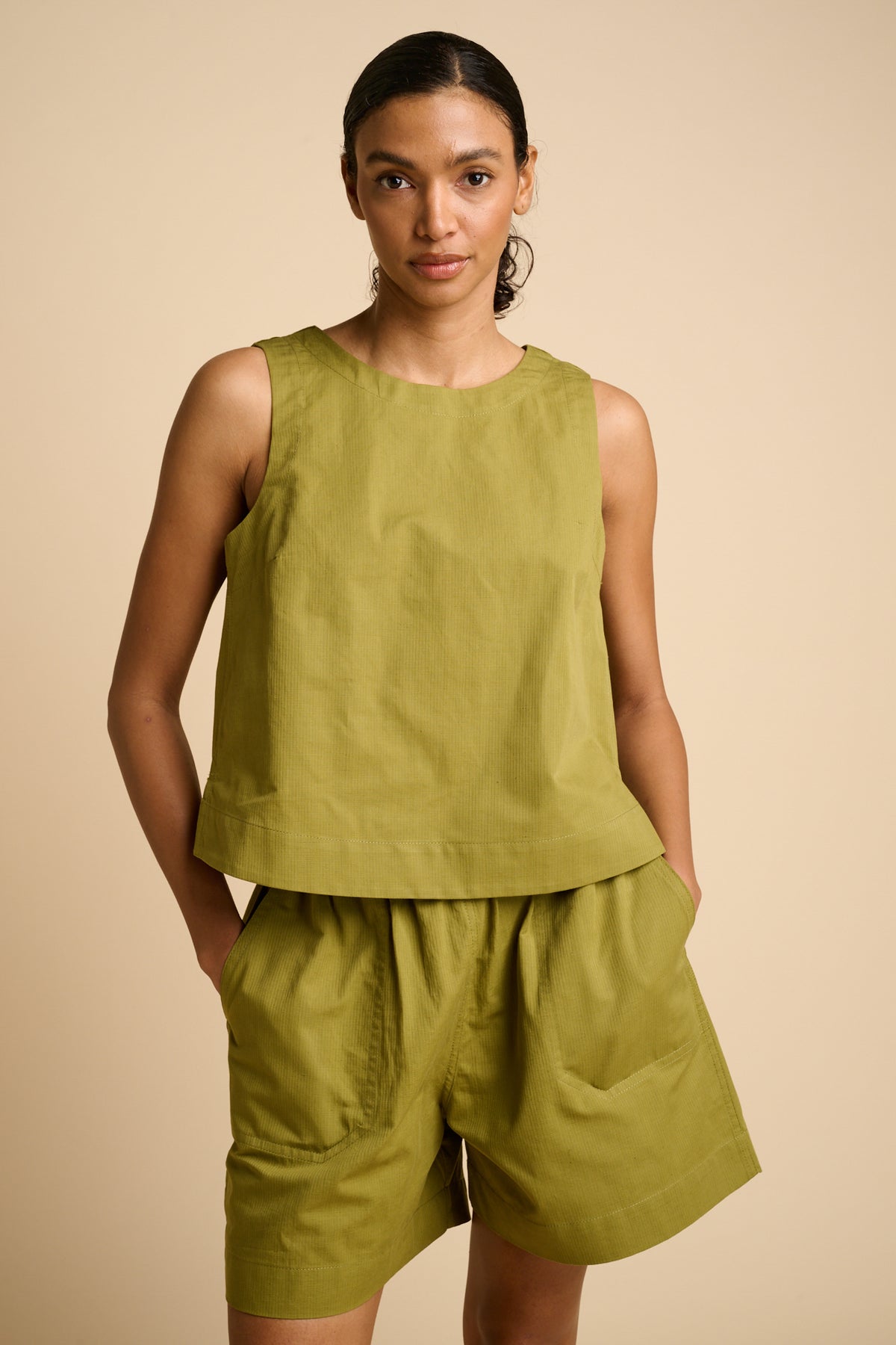 Woman wearing a green sleeveless top and shorts against a beige background.