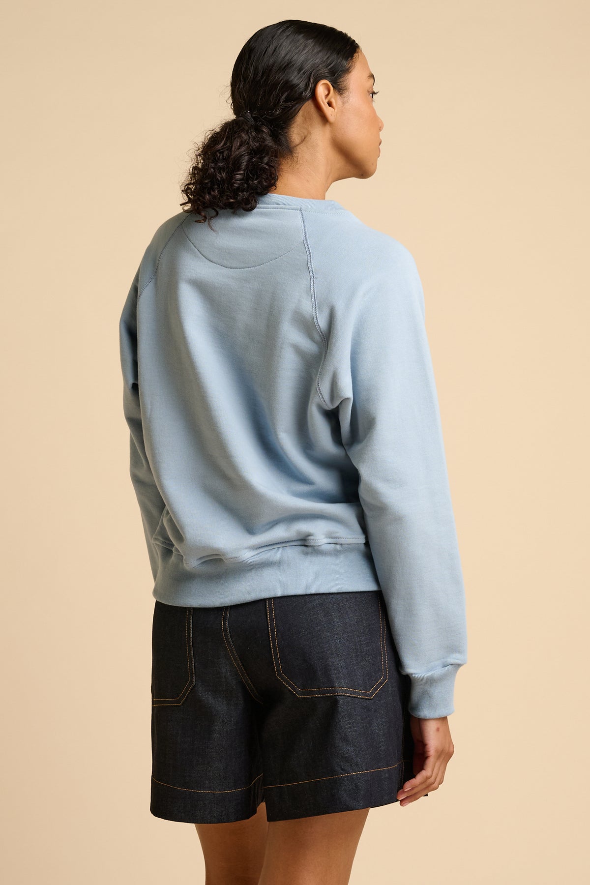 Woman wearing a sky blue sweatshirt and indigo denim shorts on a beige background.