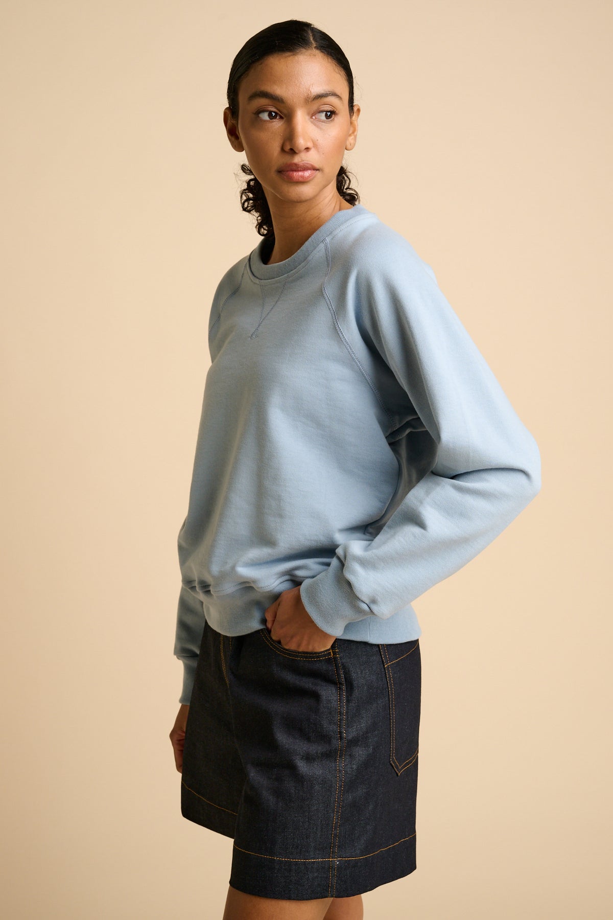 Woman wearing a sky blue sweatshirt and indigo denim shorts on a beige background.