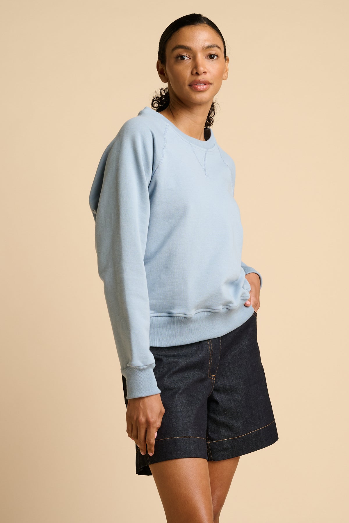 Woman wearing a sky blue sweatshirt and indigo denim shorts on a beige background.