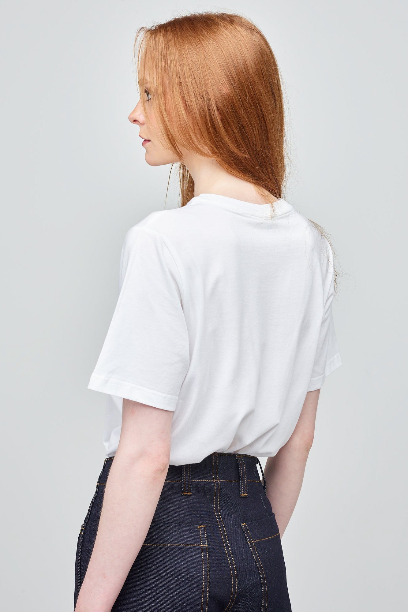 Actress Charlotte Spencer wearing a Short Sleeve white T-Shirt with branding on the chest, stood against a white brick wall