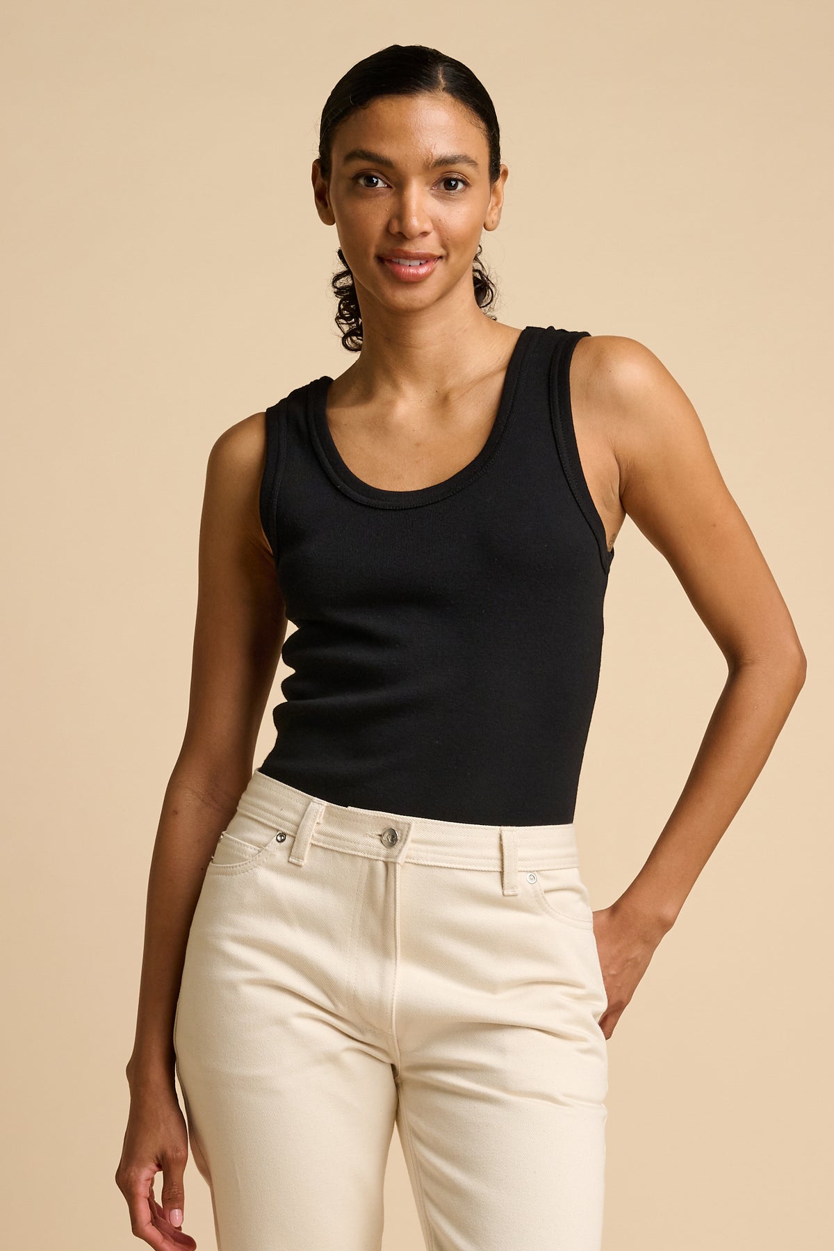 Woman wearing a black tank top and white pants against a beige background