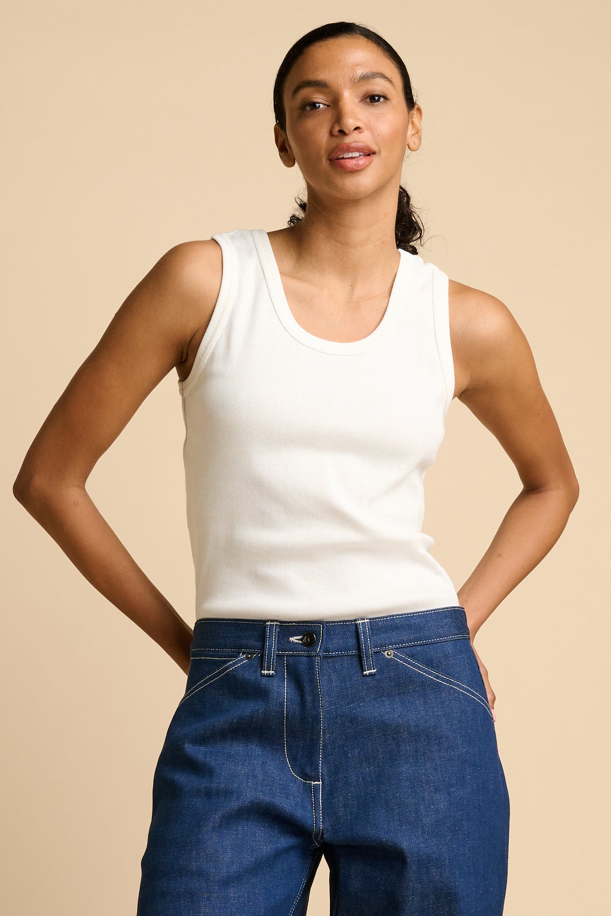 Woman wearing a white tank top and blue jeans against a beige background