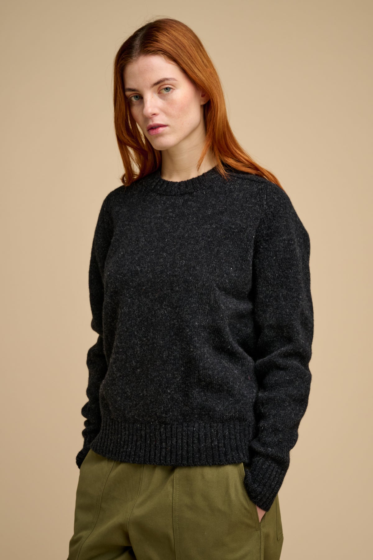 Woman wearing charcoal shetland knit jumper and green trousers against beige background.