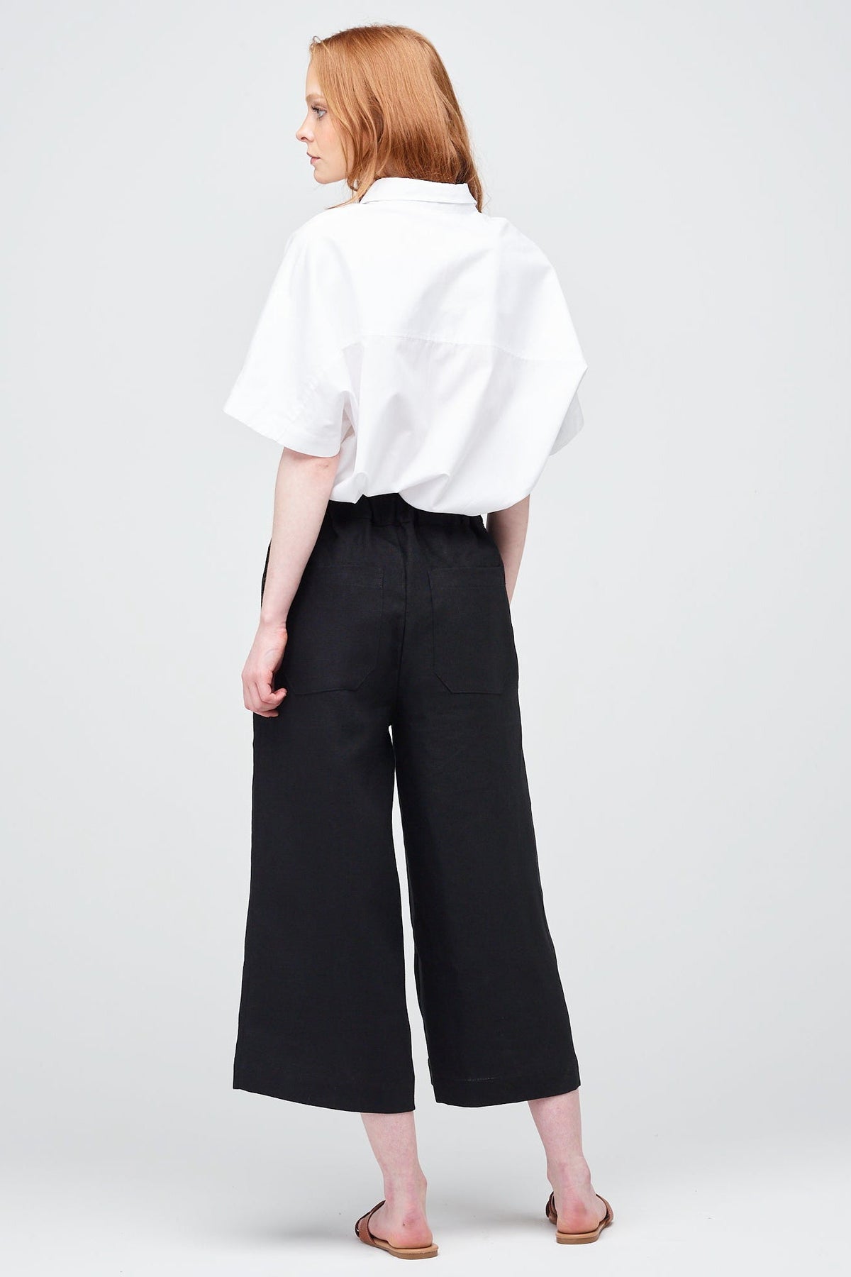 Rear shot of a young white women with ginger hair wearing short pj trouser in black linen, styled with an oversized white shirt and brown sandal.