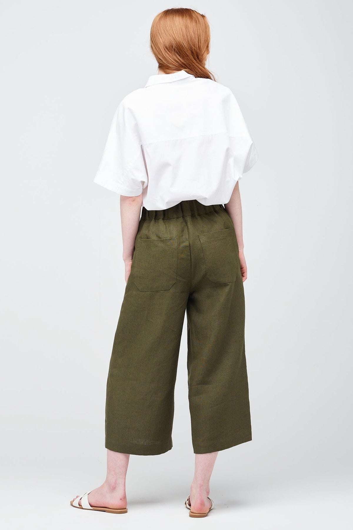 Rear shot of a young white women with ginger hair wearing short pj trouser in a khaki coloured linen, styled with an oversized white shirt and sandal.