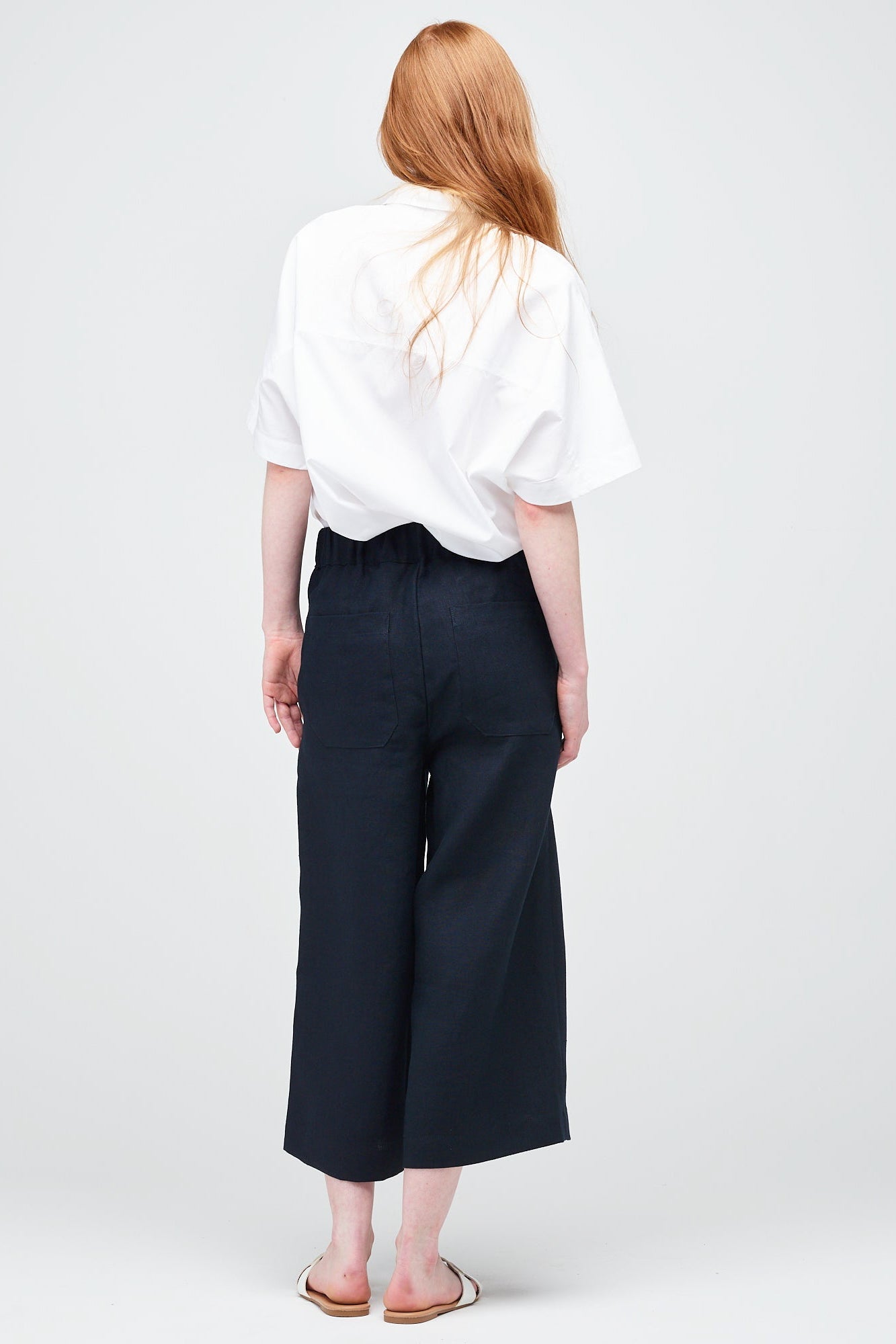 A young white women with ginger hair wearing short pj trouser in navy linen, styled with an oversized white shirt and sandal.