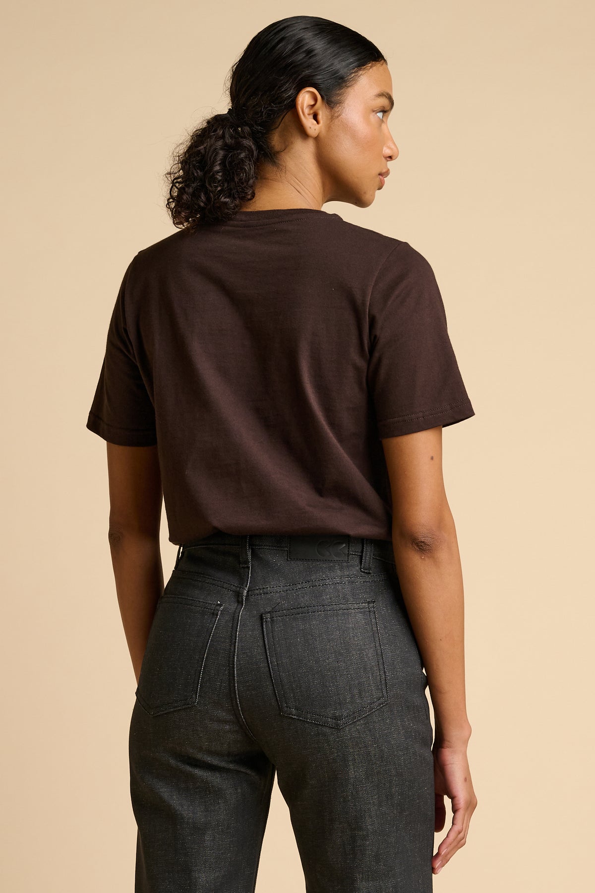 Person wearing a brown t-shirt and black jeans on a beige background.