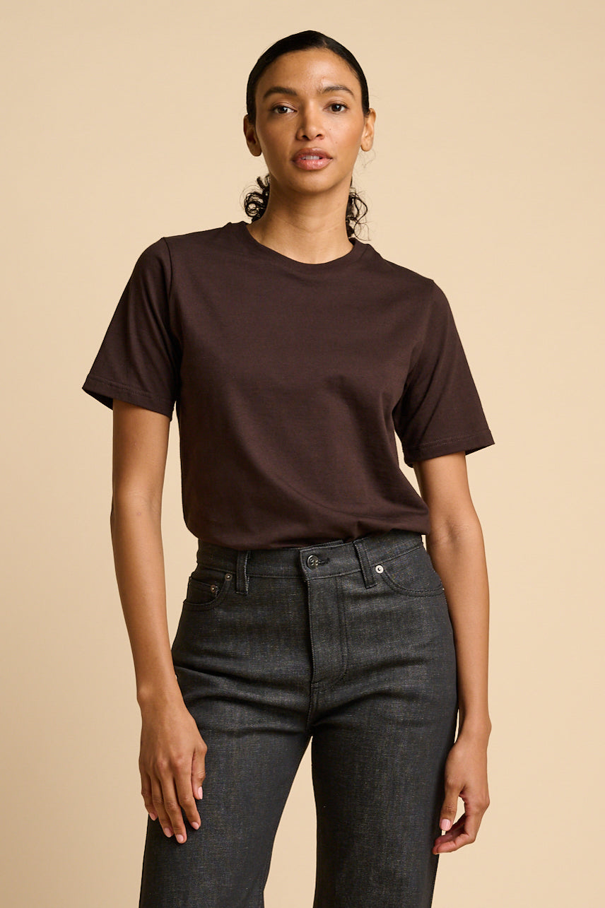 Woman wearing a brown t-shirt and black jeans against a beige background.