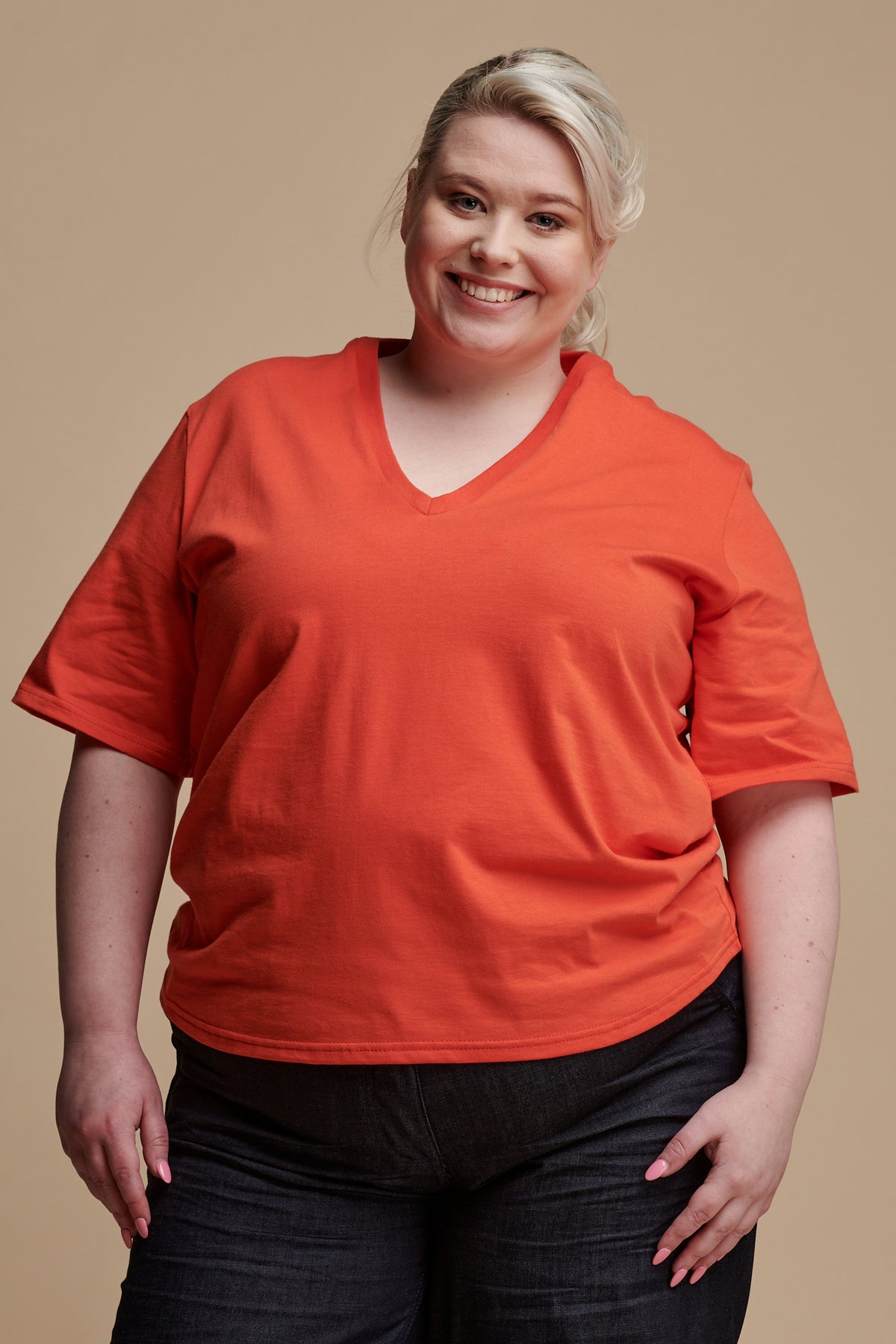 Thigh up of female with blonde hair tied back wearing short sleeve v neck t shirt in flame, paired with work trousers in denim.