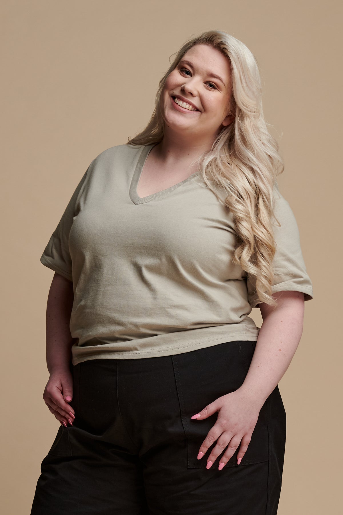 Thigh up of smiley female with long, curled blonde hair wearing short sleeve v neck t shirt in stone paired with camerawoman pants in black.