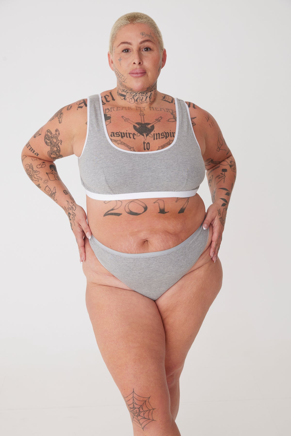 Plus sized female with short blonde hair wearing a soft jersey bra in grey with a matching low rise brief.  All from Community Clothing.